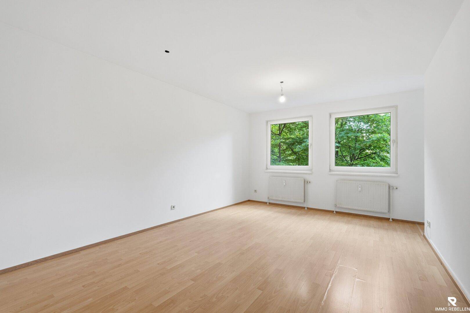 Geräumiges, leeres Zimmer mit Parkettboden und großen Fenstern mit Blick ins Grüne.
