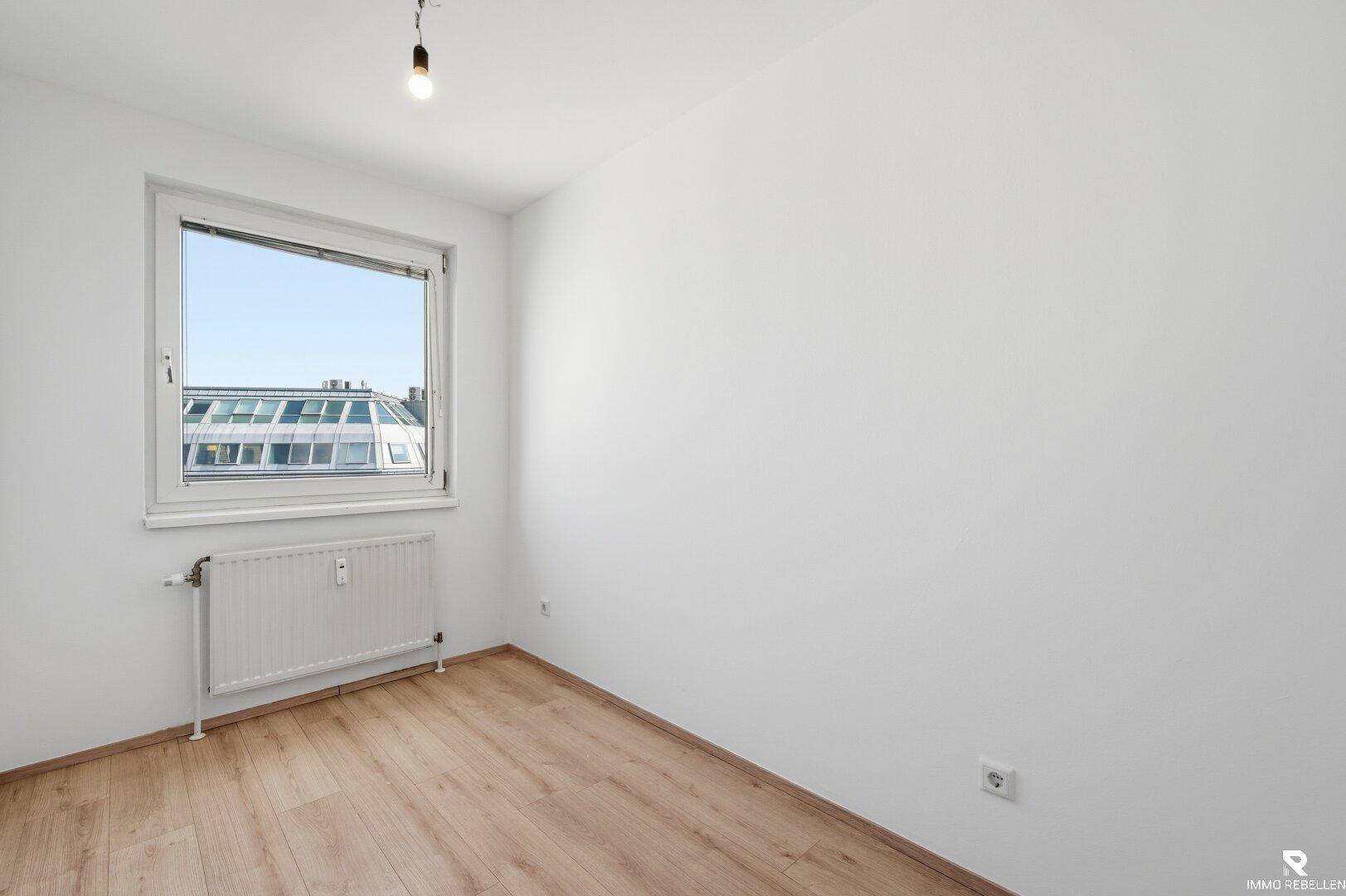 Helles, leeres Zimmer mit Laminatboden und einem Fenster mit Blick auf die Stadt.