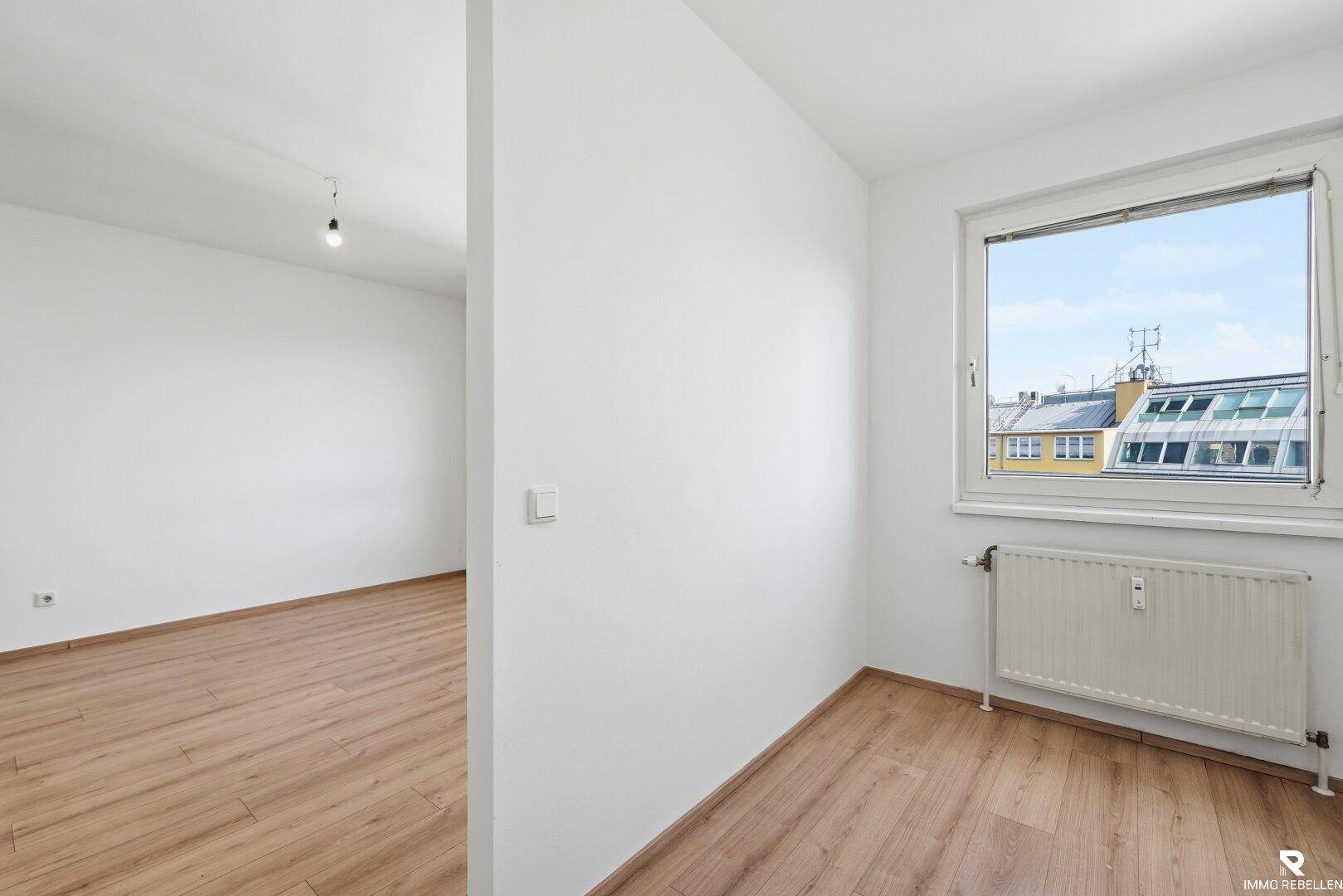 Leeres Zimmer mit Laminatboden und Fenster, das viel Tageslicht hereinlässt.
