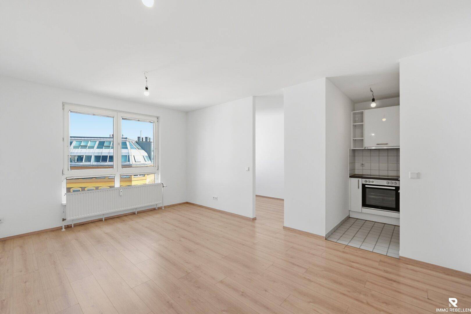 Leeres Wohnzimmer mit Laminatboden und einer kleinen, funktionalen Küchenzeile.