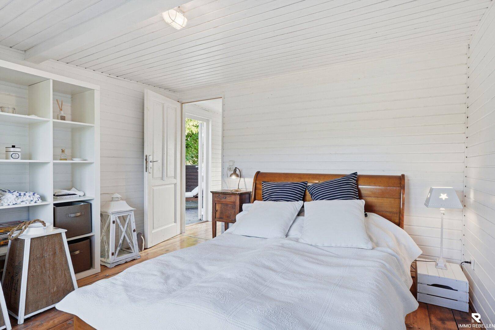 Helles Schlafzimmer mit weiß gestrichenen Holzwänden, Holzboden und Zugang nach draußen.