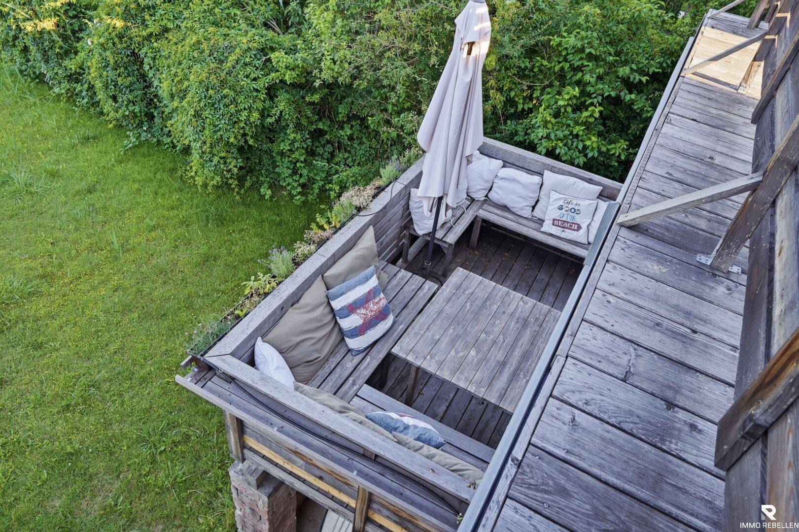Gemütliche Holzterrasse mit Sitzgelegenheiten, Kissen und Sonnenschirm, umgeben von üppigem Grün.