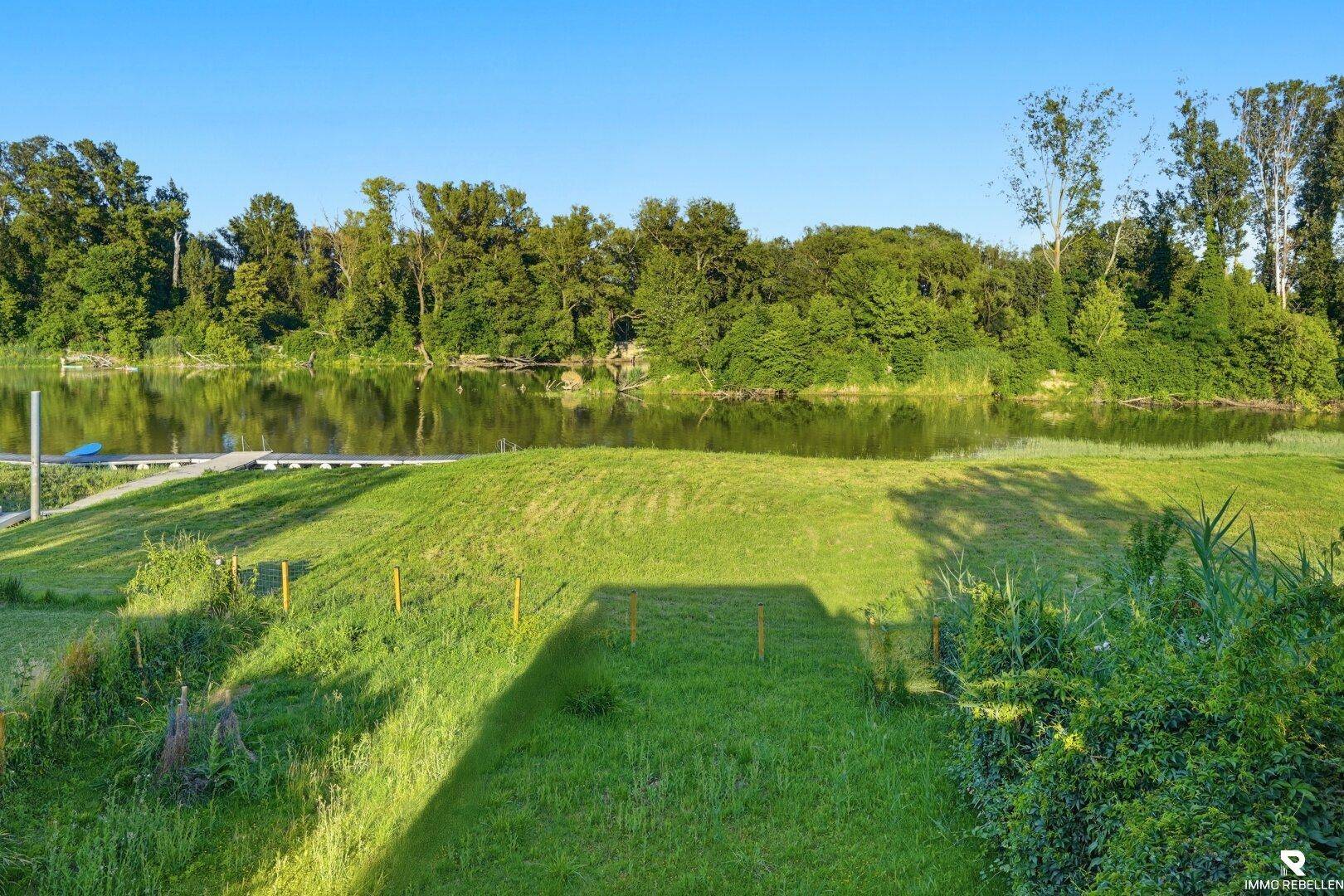 Grüne Rasenfläche mit Zugang zu einem ruhigen Fluss oder See, umgeben von Bäumen.