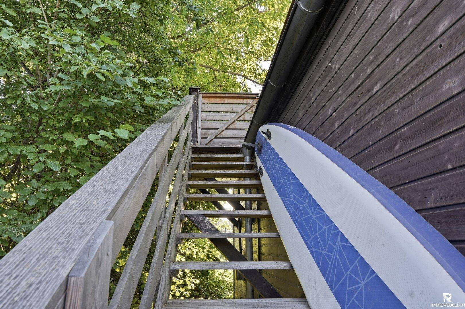 Rustikale Holztreppe im Freien, die zu einem oberen Bereich führt, mit einem Paddleboard daneben.