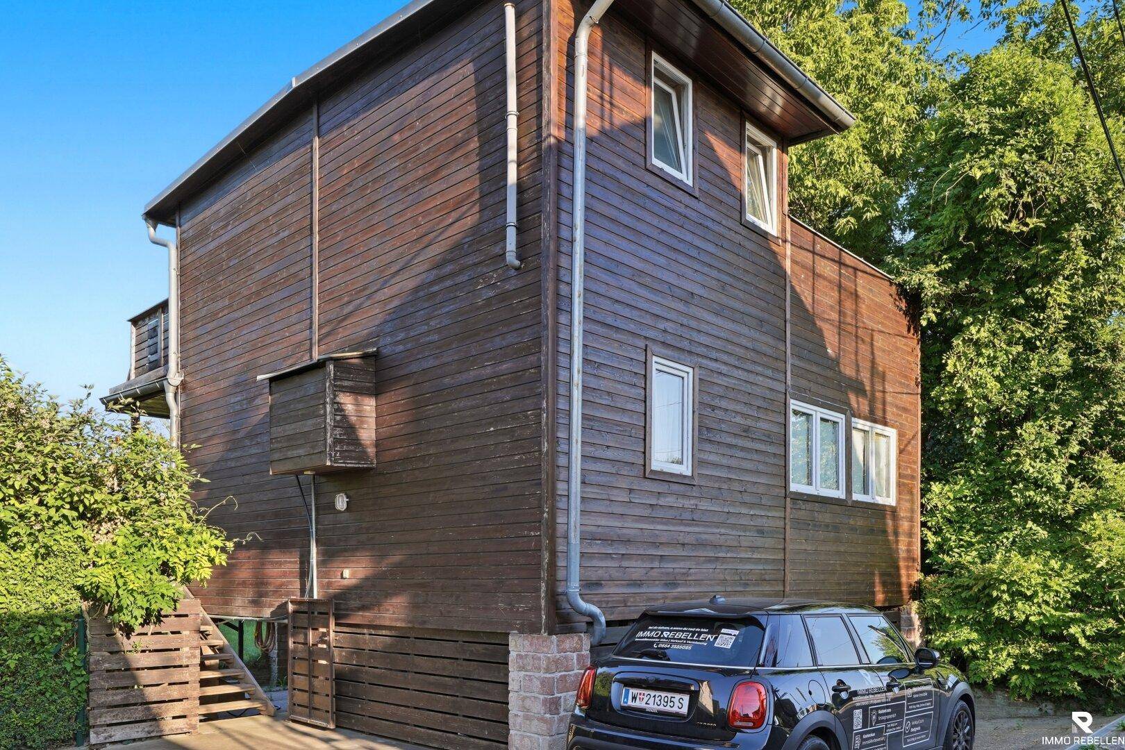 Dunkelbraunes Holzhaus mit mehreren Fenstern und einem geparkten Auto davor, umgeben von Bäumen.