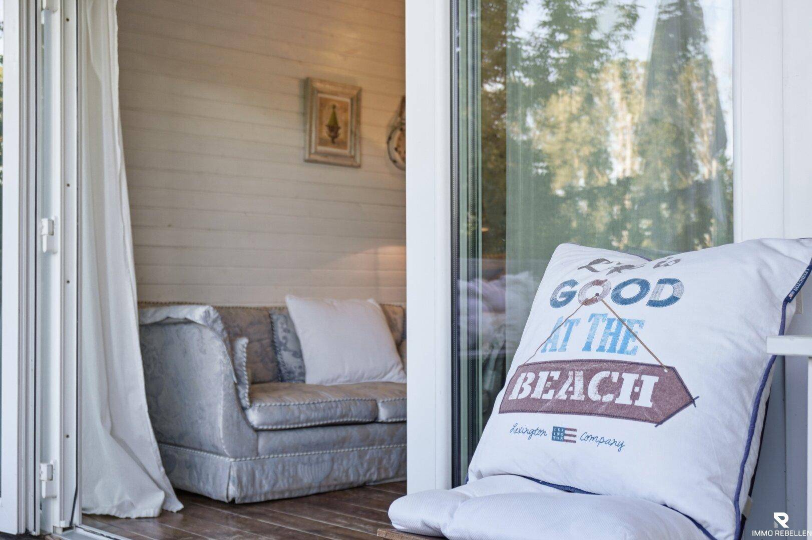 Heller Innenraum mit weiß getäfelten Wänden und einem bequemen Sofa, gesehen durch eine Glastür.