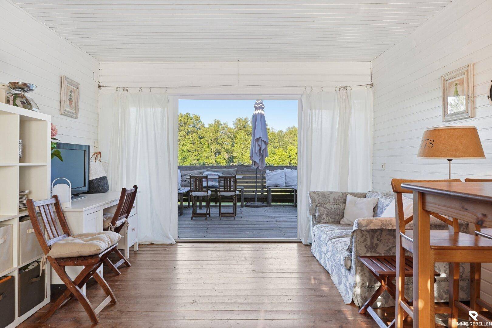 Helles Wohnzimmer mit Holzboden, weiß getäfelten Wänden und direktem Zugang zur Terrasse.
