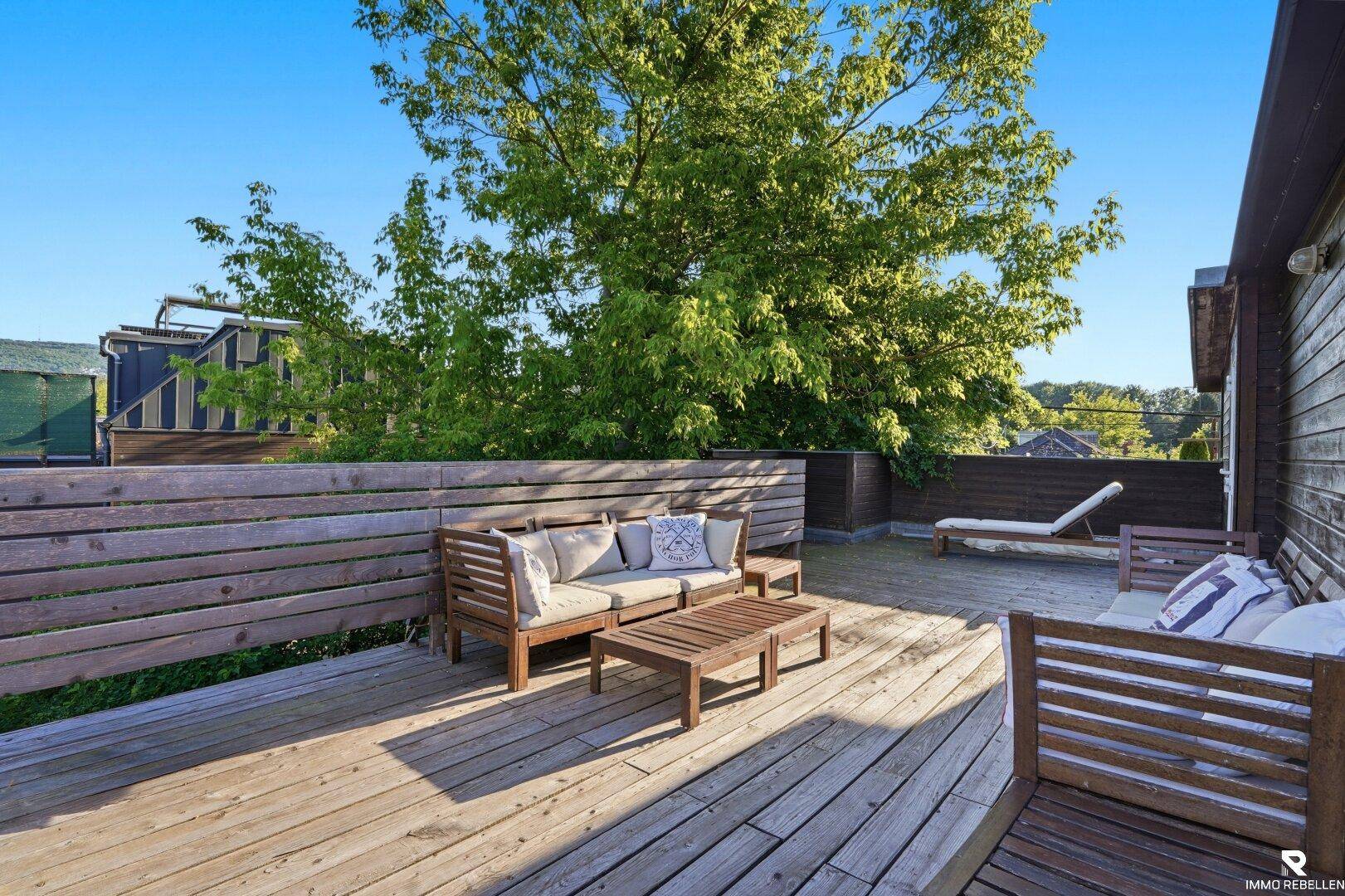 Sonnige Holzterrasse mit modernen Lounge-Möbeln und viel Grün im Hintergrund.