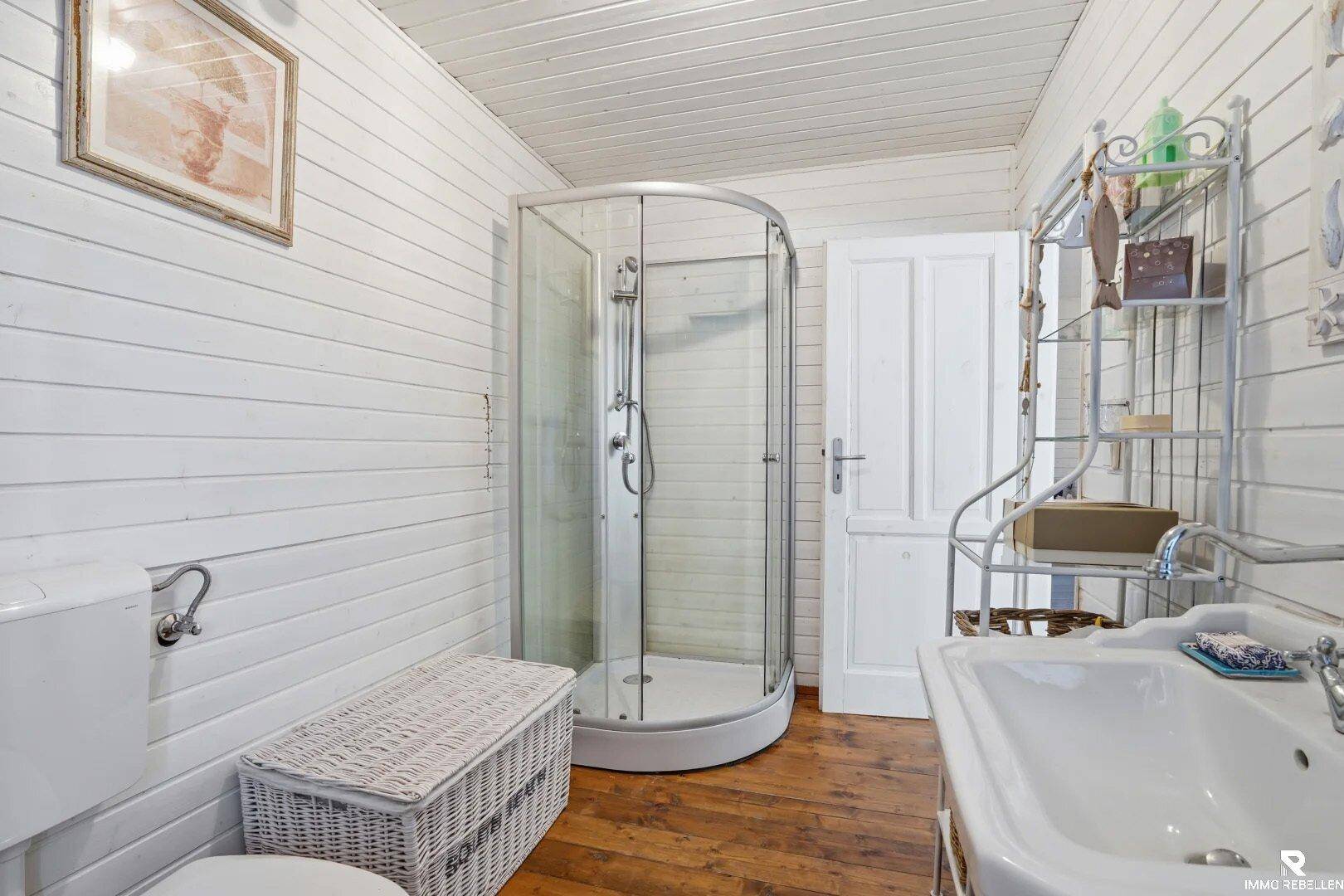 Helles Badezimmer mit Duschkabine, Waschbecken und weißen Holzpaneelen.