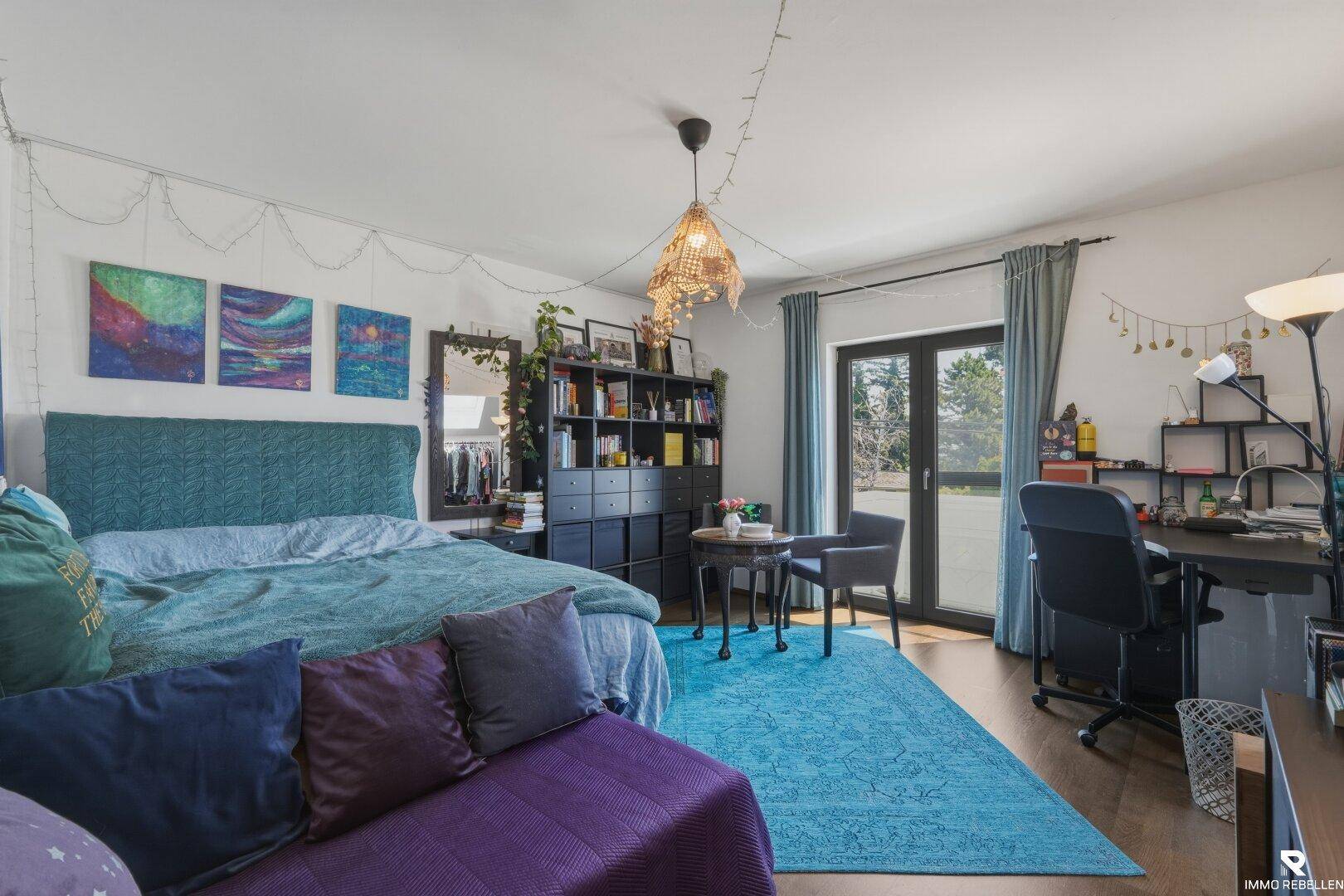 Helles Schlafzimmer mit Balkonzugang, Schreibtisch und farbenfroher Dekoration.