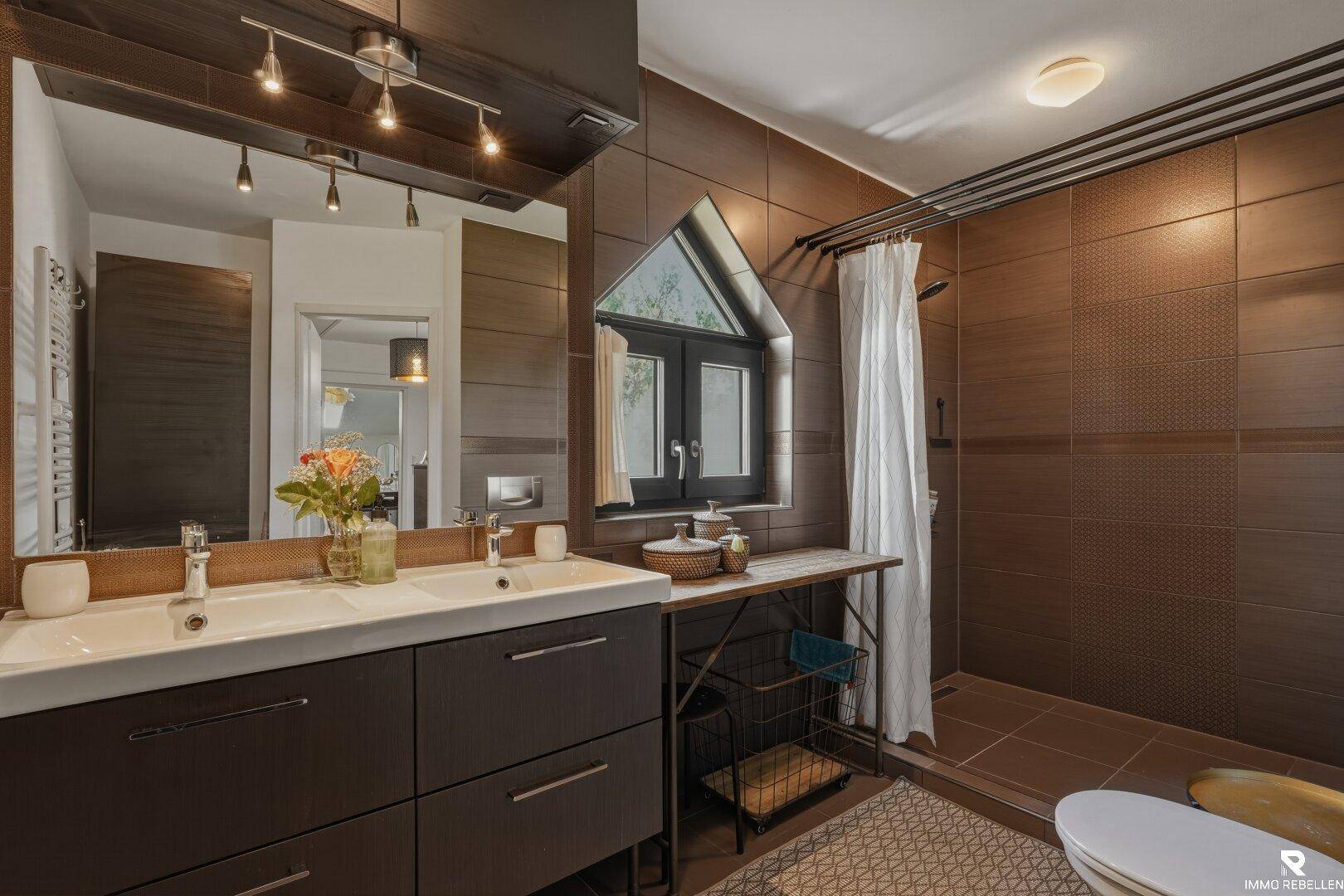 Modernes Badezimmer mit Doppelwaschbecken, braunen Fliesen und Duschbereich.
