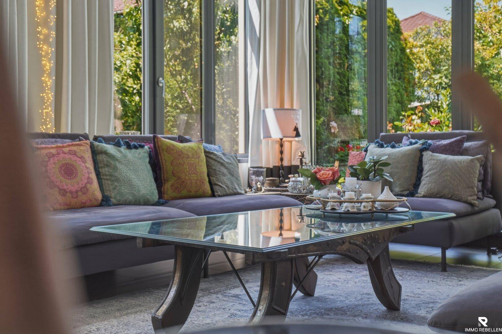 Gemütliches Wohnzimmer mit Sofa und Glastisch, umgeben von großen Fenstern und viel Tageslicht.