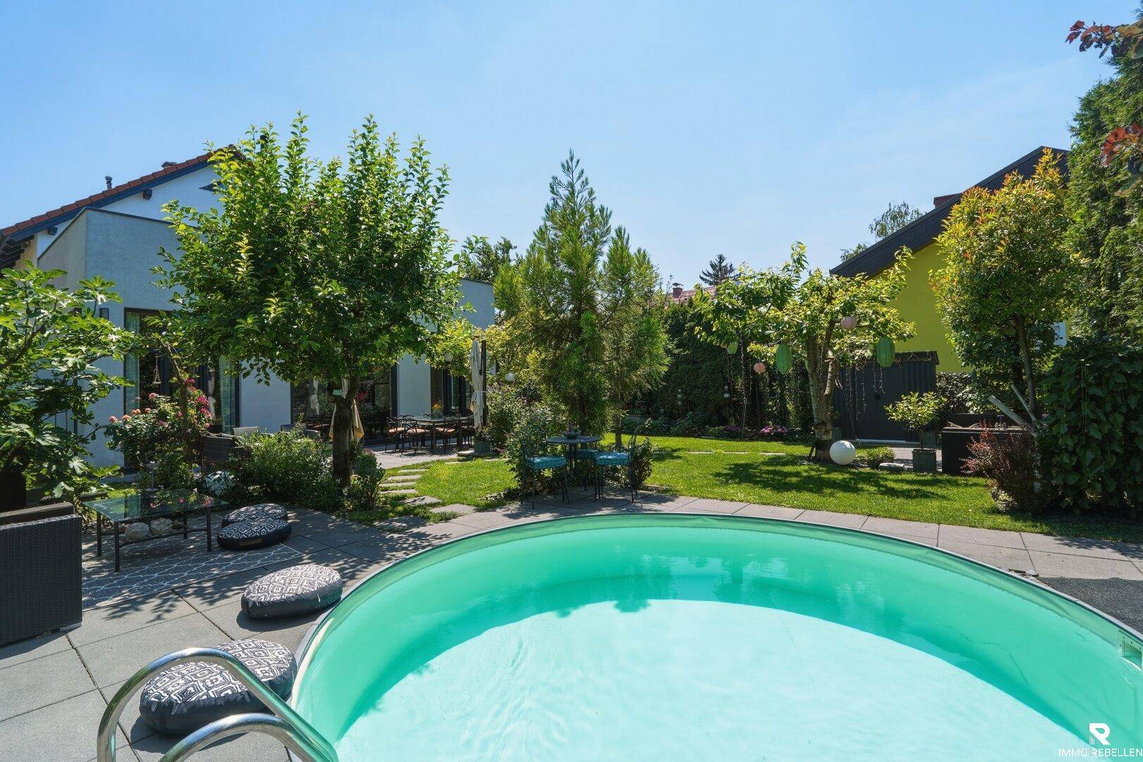 Sonniger Garten mit einem runden Pool, umgeben von Rasenflächen und üppiger Vegetation.