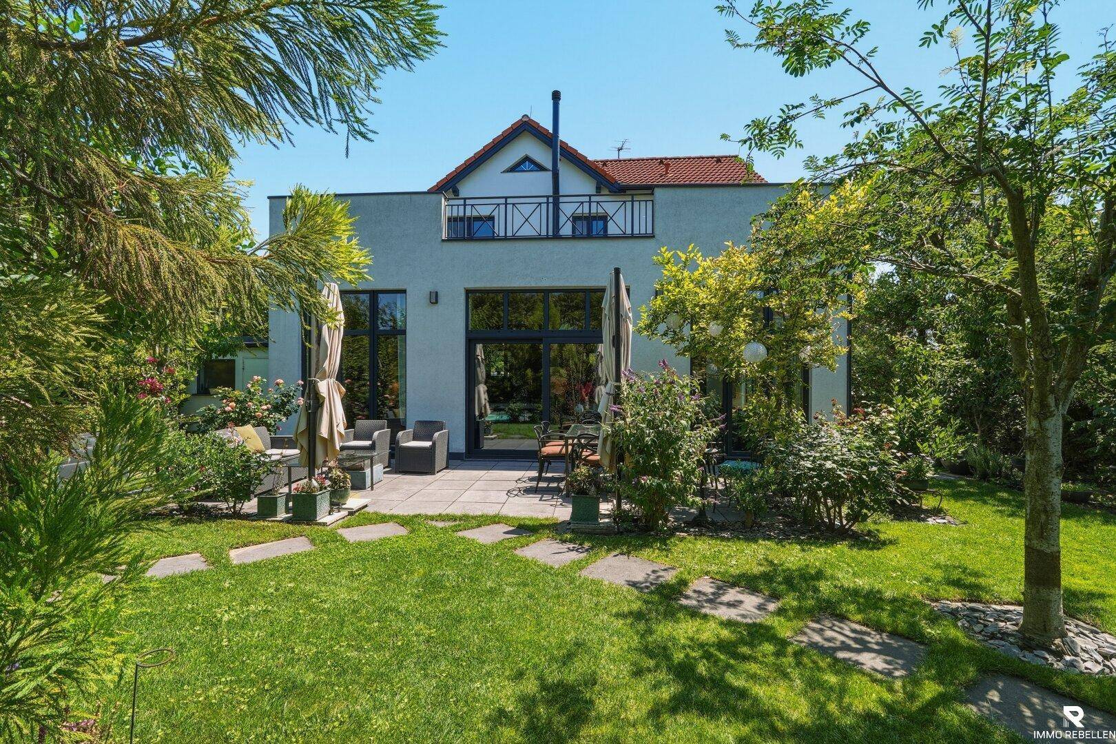 Rückseite des Hauses mit großer Terrasse, Rasenfläche und üppiger Bepflanzung im Garten.