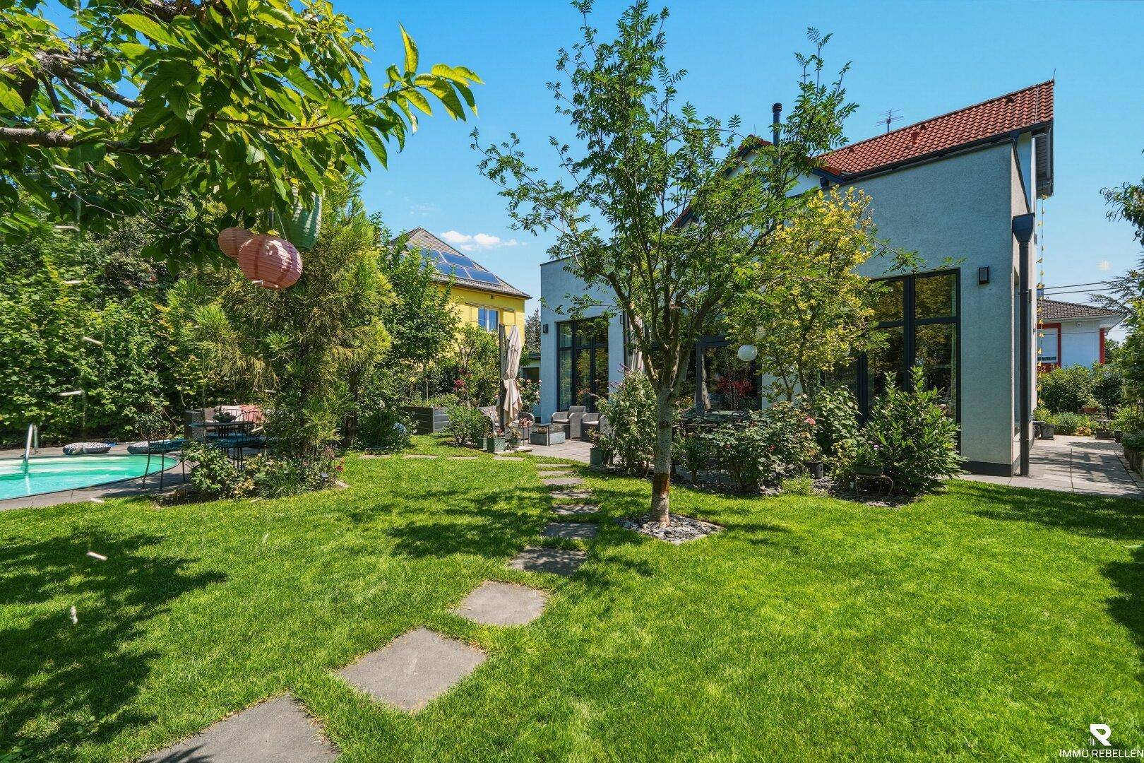 Gepflegter Garten mit Rasen, Bäumen und einem Pool im Hintergrund, angrenzend an die Hausfassade.