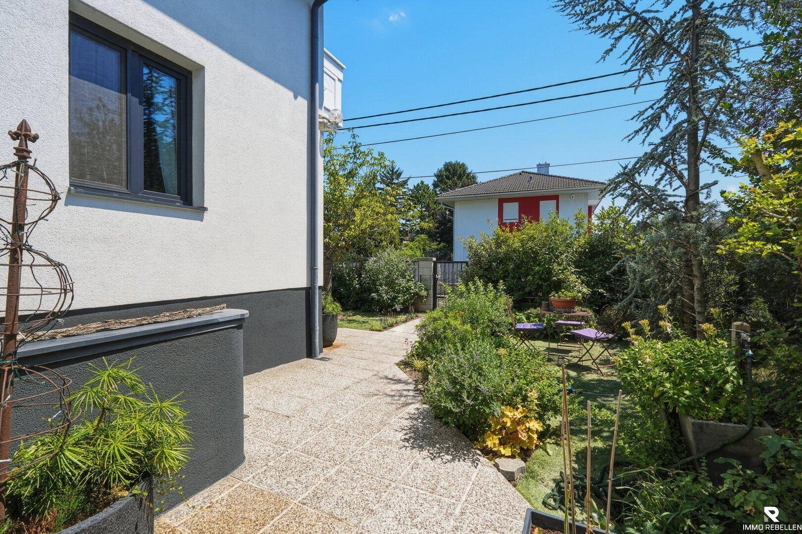 Seitlicher Weg des Hauses, der zu einem grünen Gartenbereich mit weiteren Sitzgelegenheiten führt.