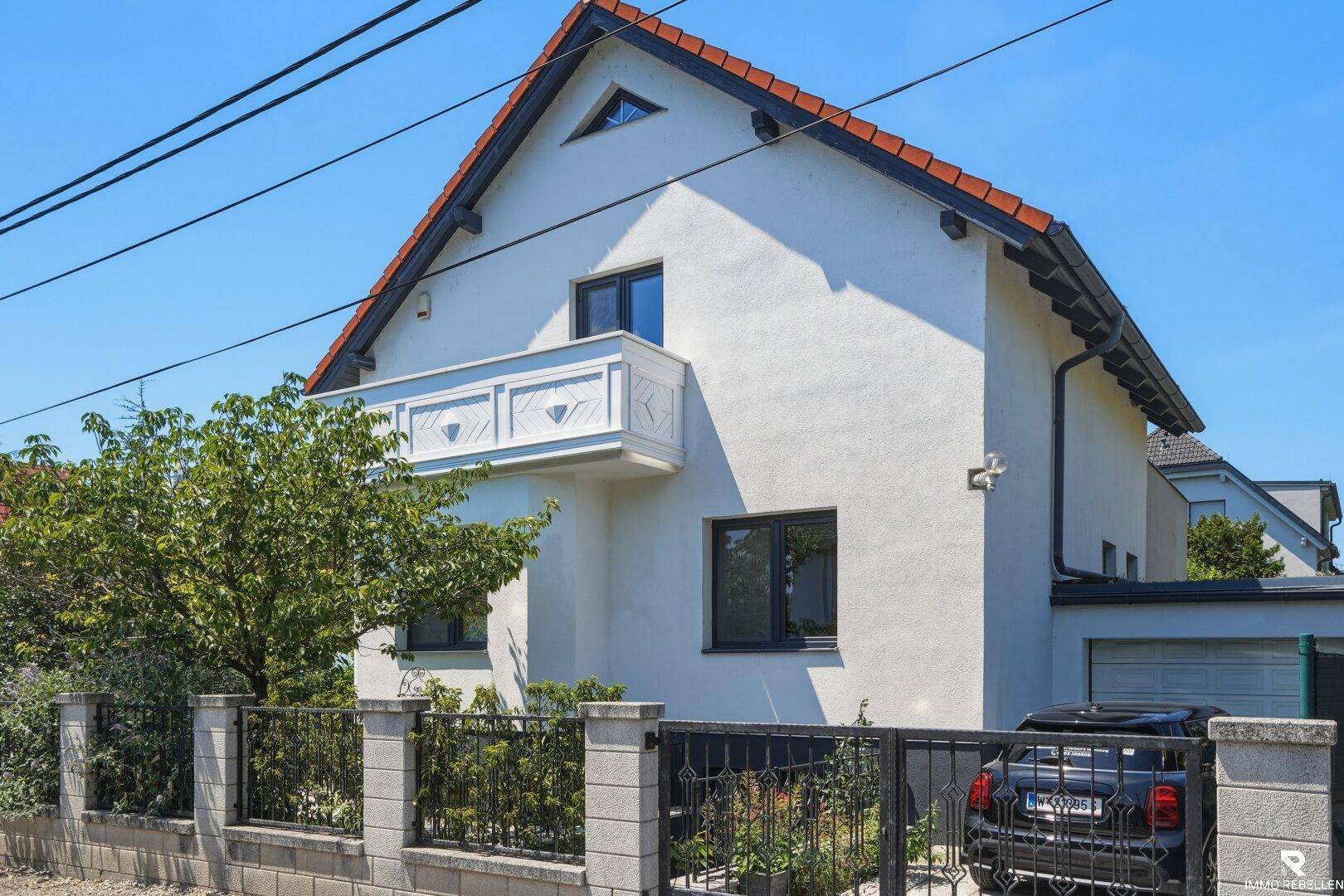 Seitenansicht des Hauses mit Balkon, Garage und gepflegtem Vorgarten unter blauem Himmel.
