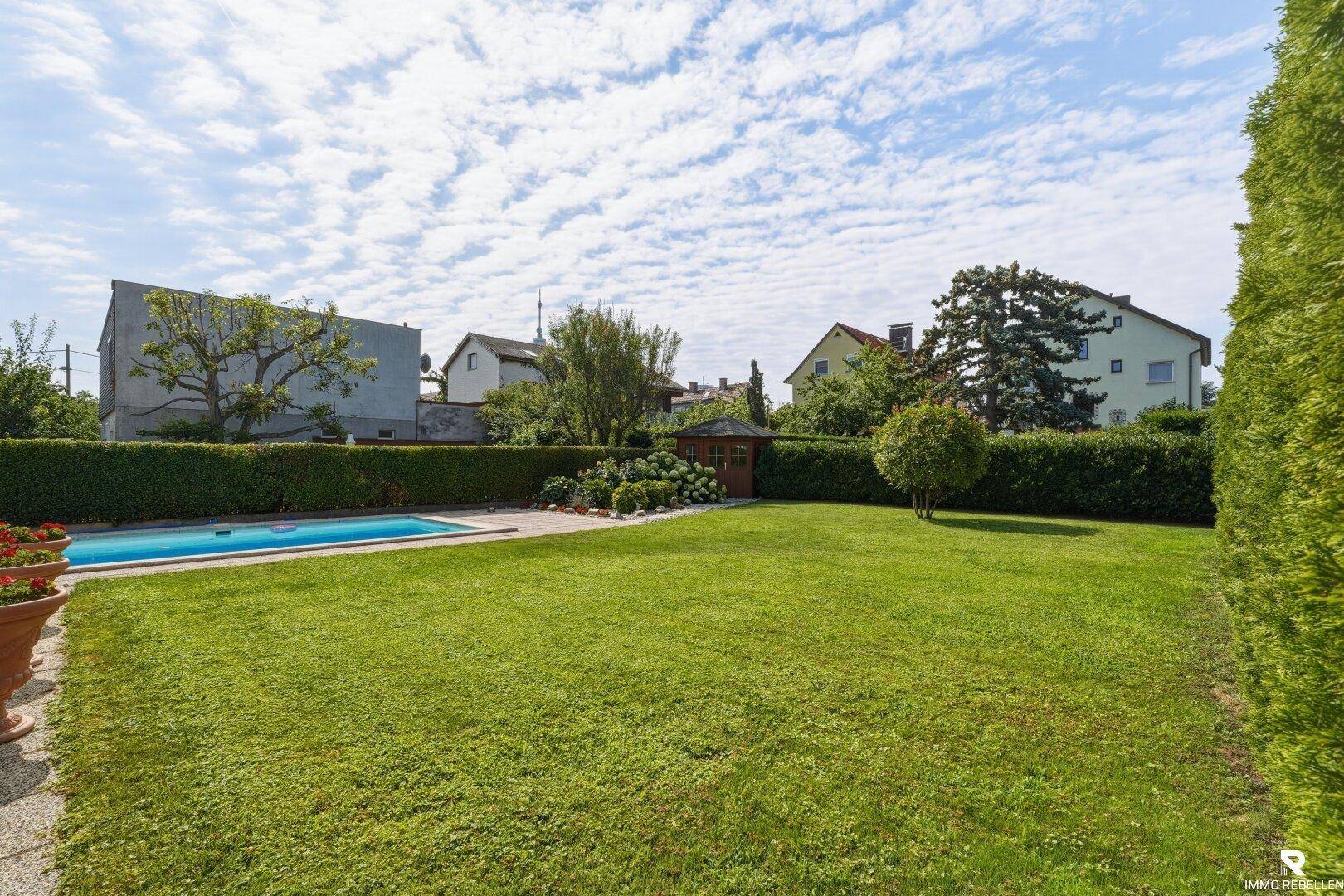 Weitläufiger Gartenbereich mit Pool, großer Rasenfläche und einem kleinen Gartenhaus unter blauem Himmel.