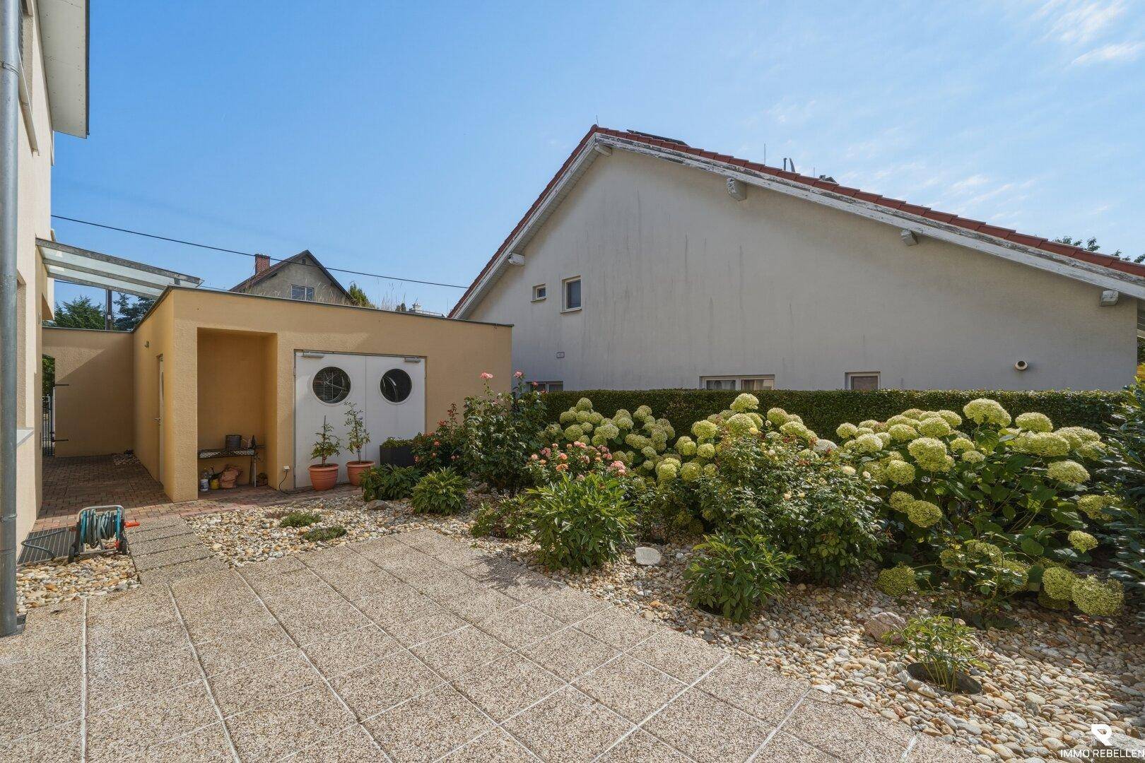 Gepflegter Gartenbereich mit Steinplatten, Ziersträuchern und einem kleinen Nebengebäude.