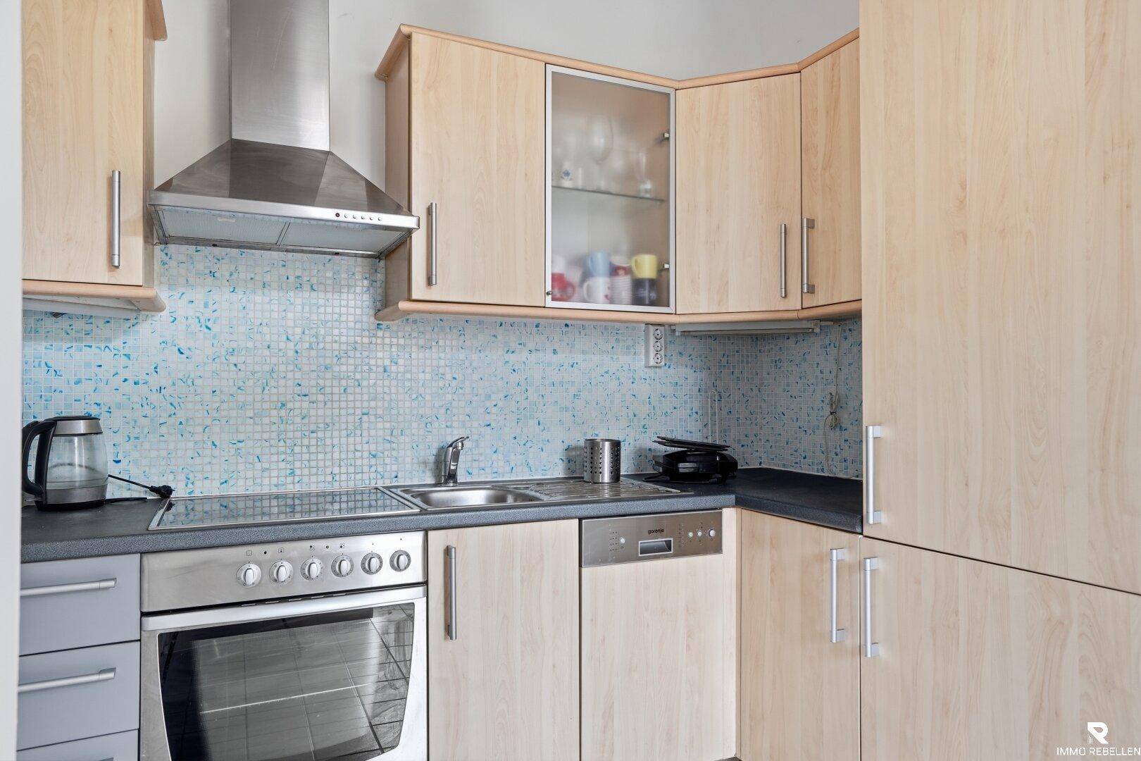 Funktionale Küche mit hellen Holzschränken, Edelstahlgeräten und blauen Mosaikfliesen.