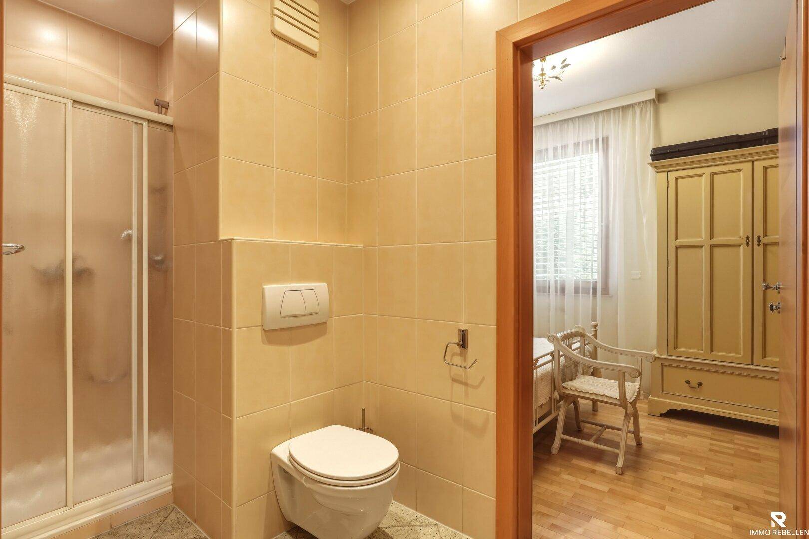 Kleines Badezimmer mit beigen Fliesen, WC und Duschkabine, angrenzend an ein Schlafzimmer.