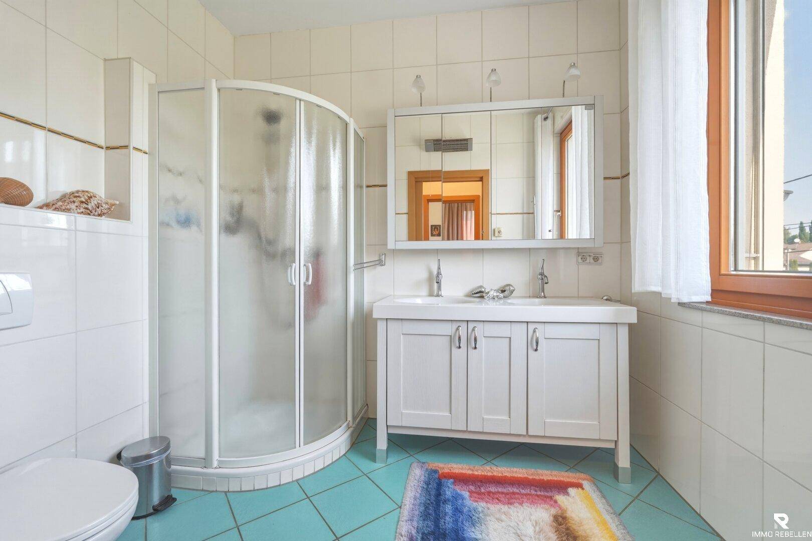 Badezimmer mit Duschkabine, Doppelwaschbecken, Spiegelschrank und Fenster für natürliche Belüftung.