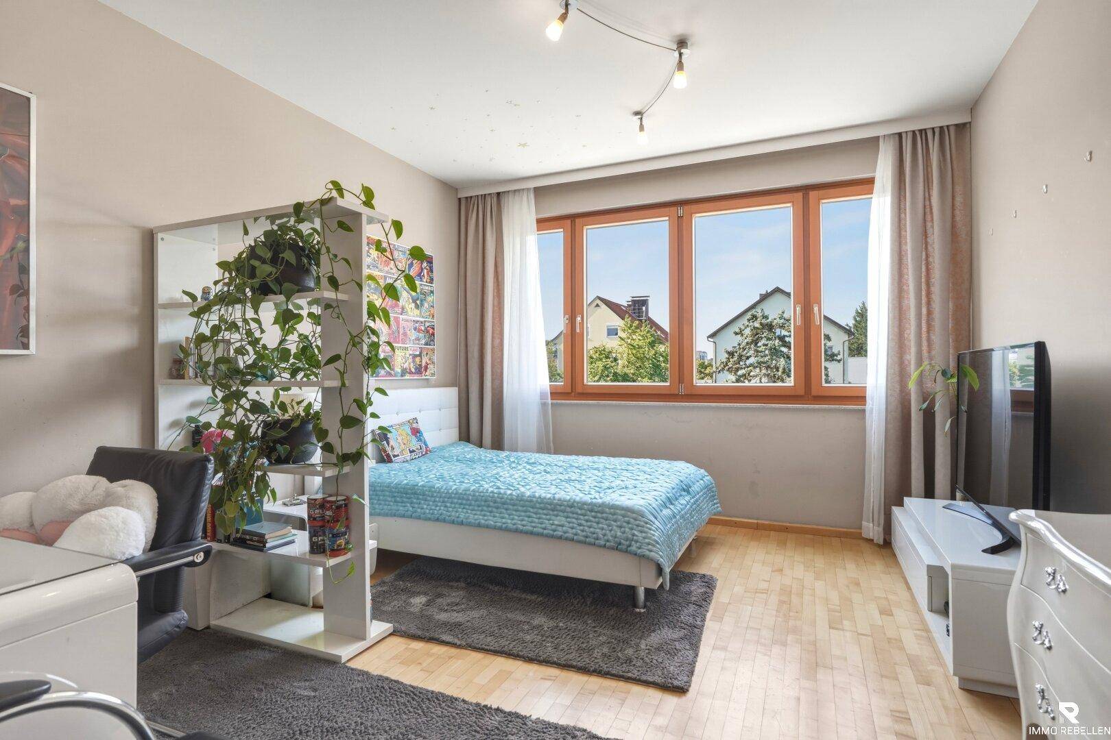 Helles Schlafzimmer mit Holzboden, großem Fenster und einem Raumteiler mit Pflanzen für Privatsphäre.