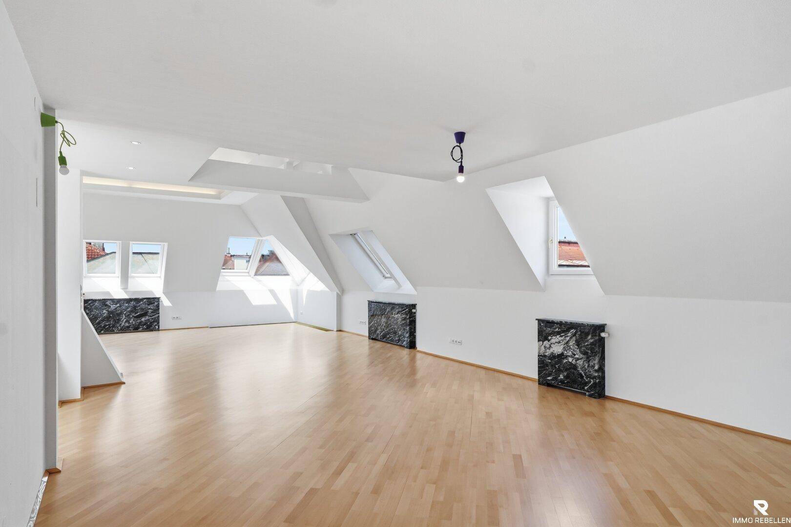 Heller, leerer Wohnraum mit Holzboden, Dachschrägen und mehreren Dachfenstern.