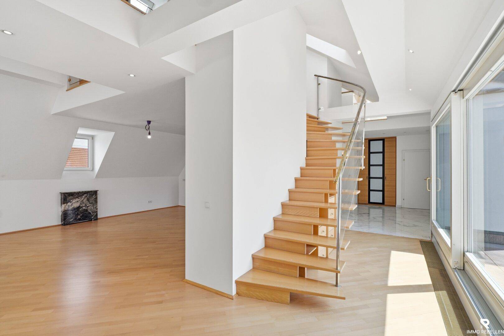 Heller Eingangsbereich mit Holztreppe, Glasgeländer und großen Fenstern, die viel Licht hereinlassen.