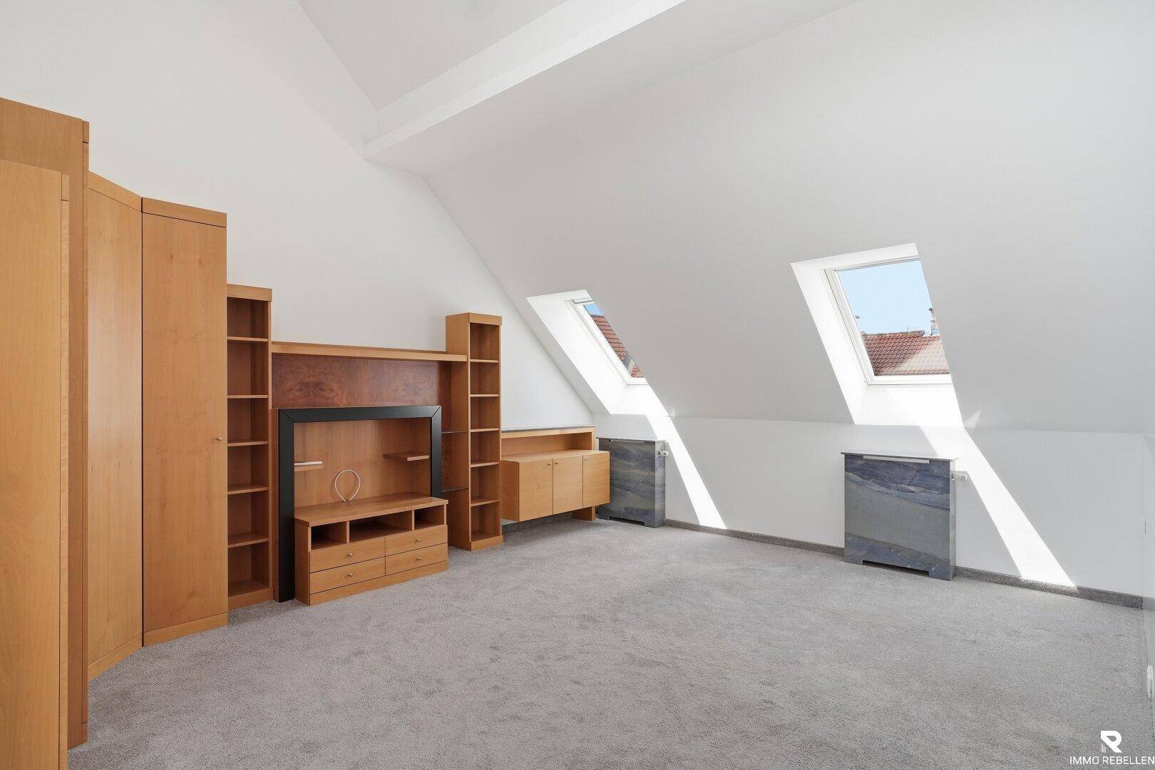 Heller Dachbodenraum mit Teppichboden, Holzmöbeln und zwei Dachfenstern.