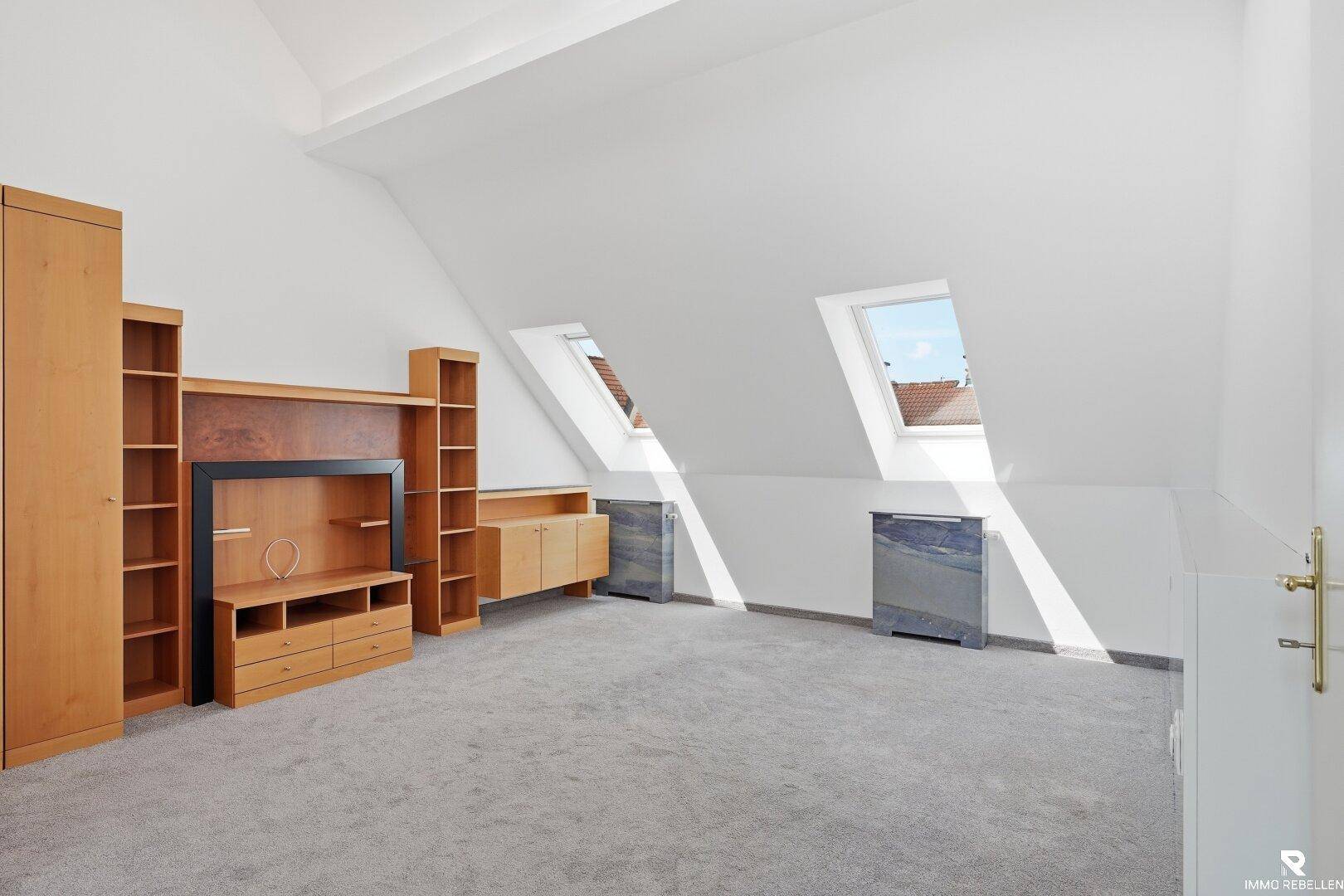 Heller Dachbodenraum mit Teppichboden, Holzmöbeln und zwei Dachfenstern.
