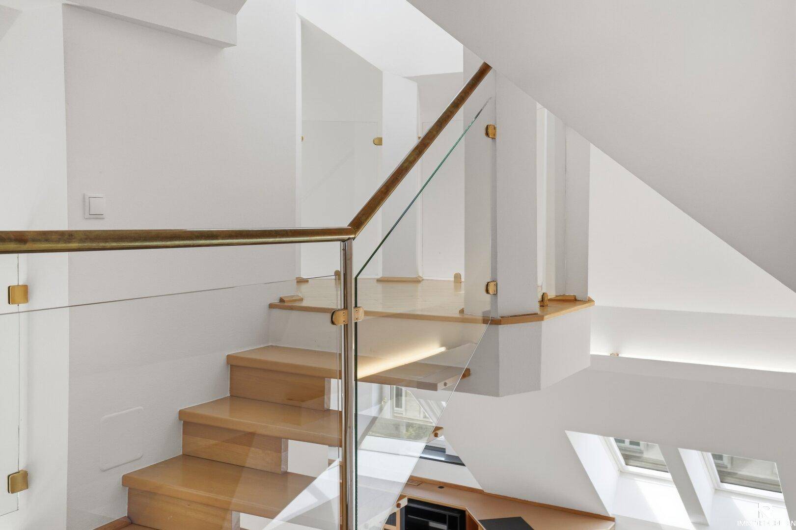 Moderne Holztreppe mit Glasgeländer und indirekter Beleuchtung in einem hellen Treppenhaus.