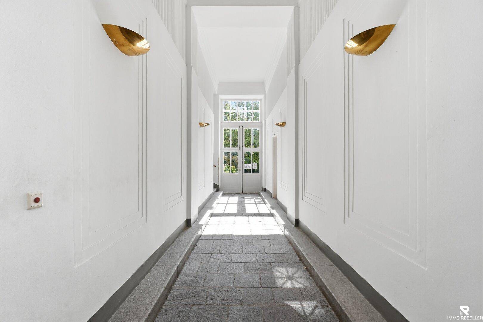 Langer, heller Flur mit hohen Decken, modernen Wandleuchten und Steinboden, der zu einer großen Fenstertür führt.