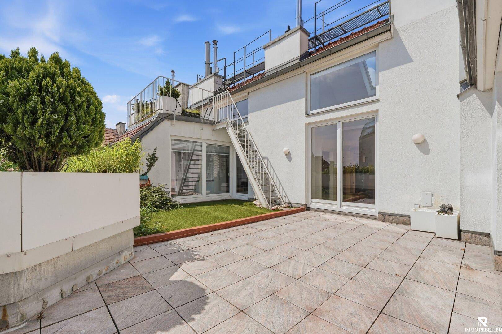Geräumige Dachterrasse mit Fliesenboden und Zugang zu einem kleinen Rasenbereich unter blauem Himmel.
