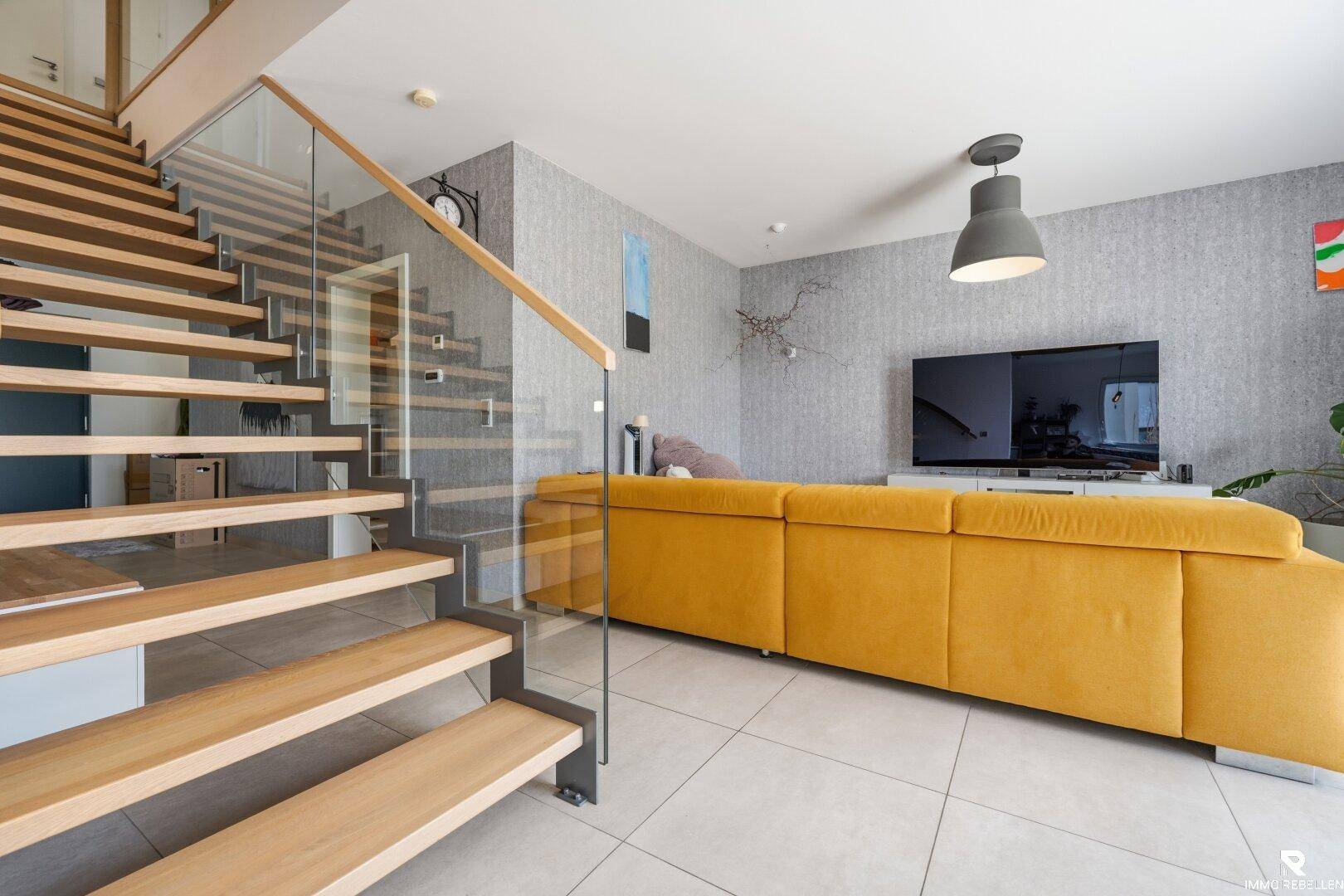 Helles Wohnzimmer mit gelbem Sofa, Fliesenboden und moderner Holztreppe mit Glasgeländer.