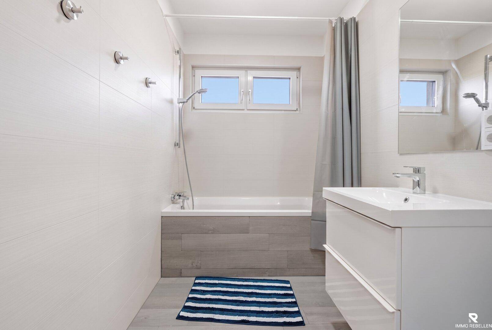 Modernes Badezimmer mit Badewanne, Waschtisch und Fenster, in hellen Farbtönen gehalten.