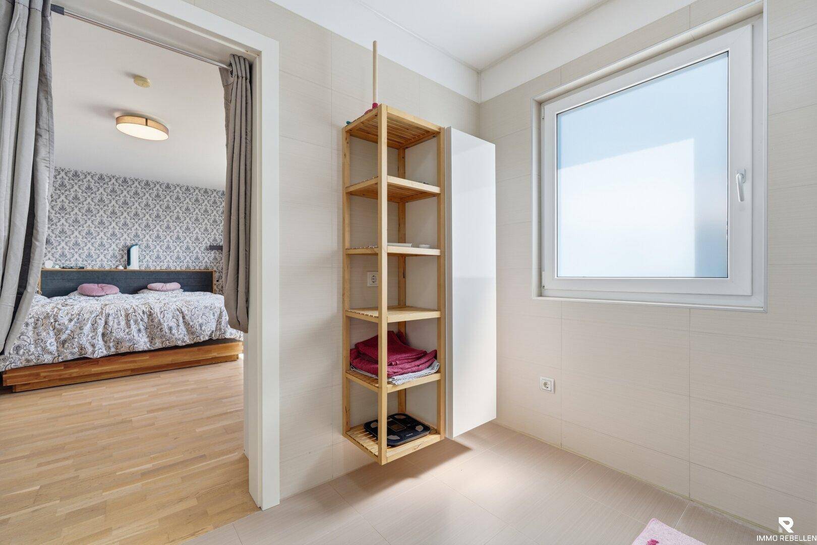 Heller Bereich vor dem Badezimmer mit Holzregal und Blick ins Schlafzimmer.