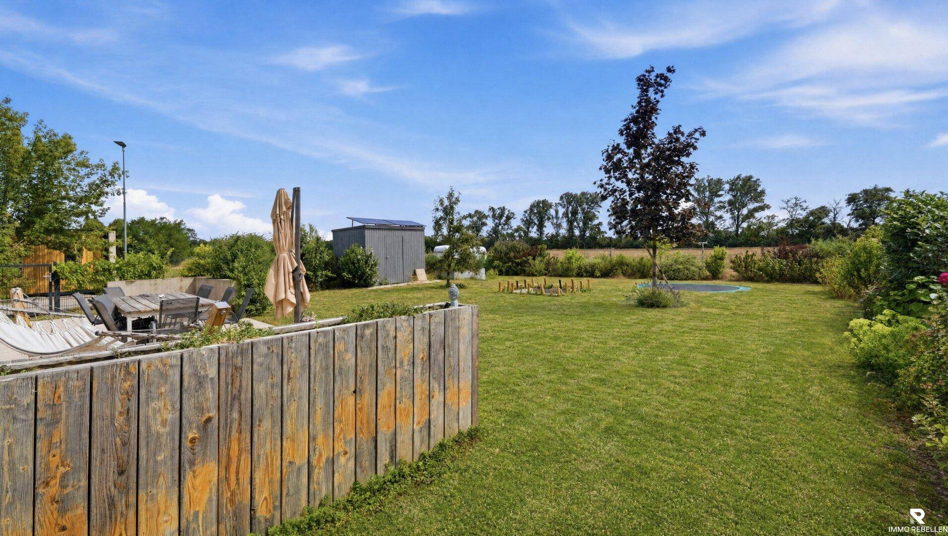 Weitläufiger Garten mit Rasenfläche, Trampolin und Gartenhaus unter blauem Himmel.