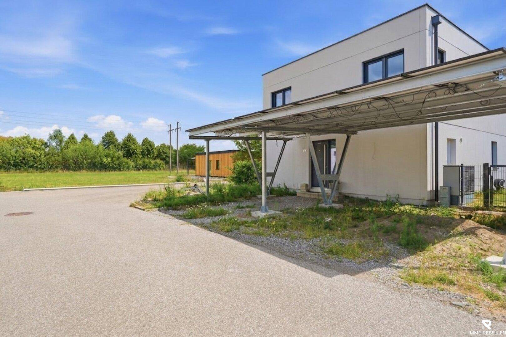 Modernes Haus mit Carport und Photovoltaikanlage, umgeben von Grünflächen.