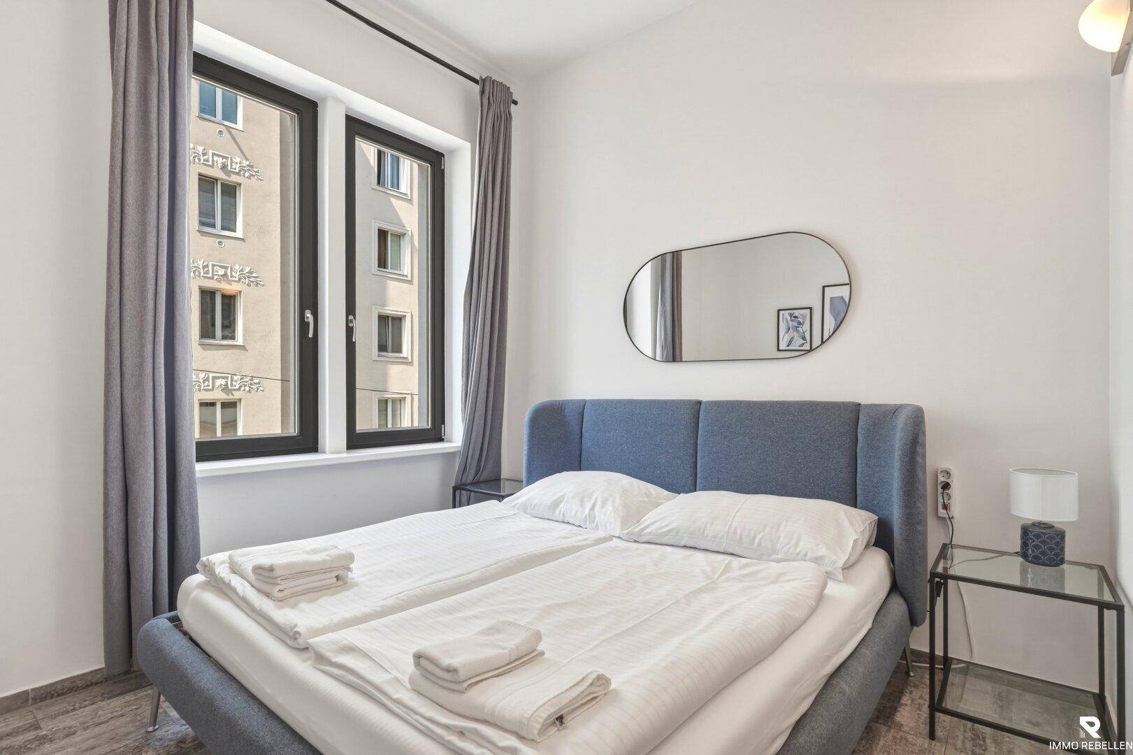 Helles Schlafzimmer mit blauem Polsterbett, Nachttisch und Blick auf die umliegenden Gebäude.