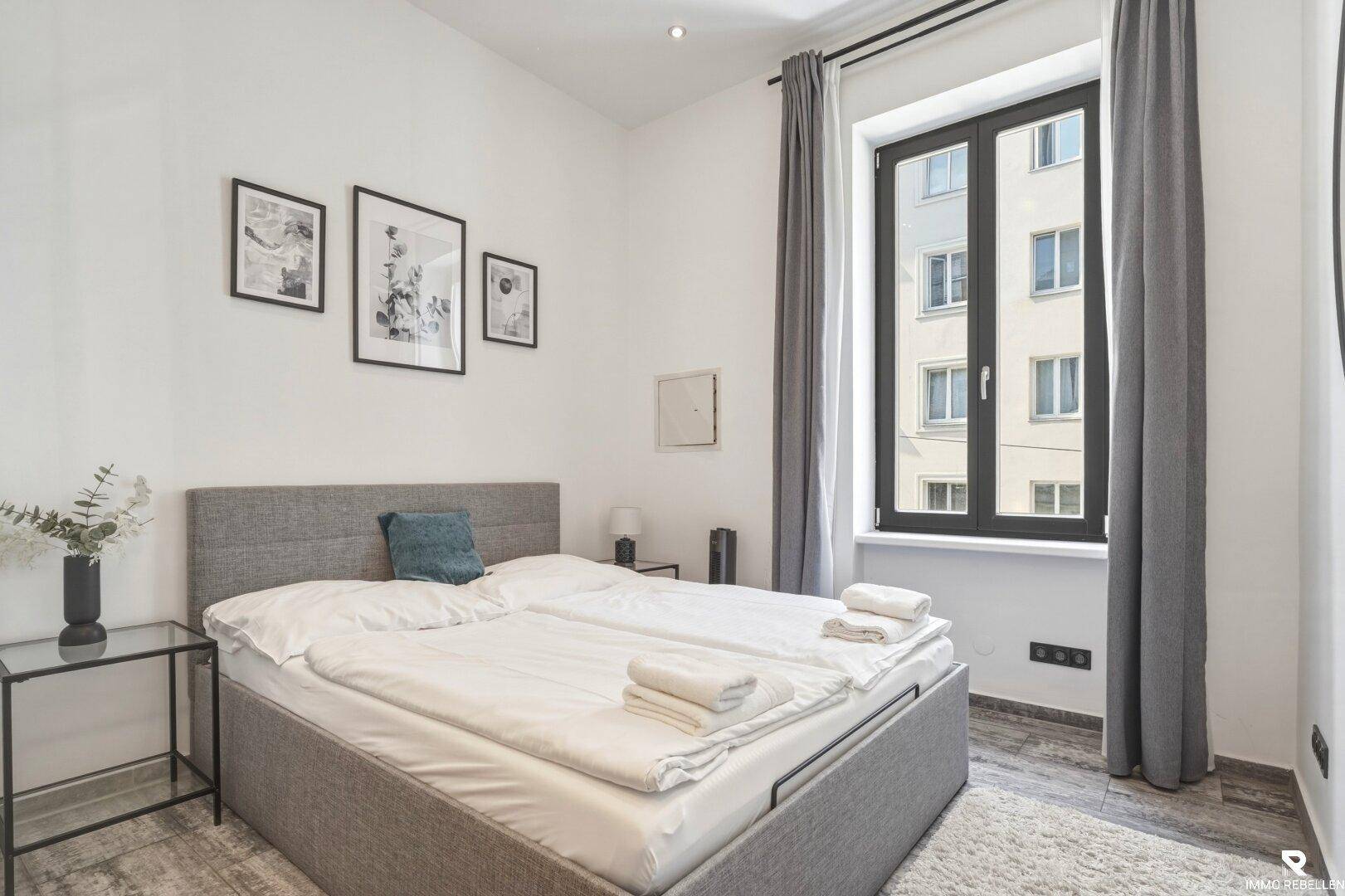 Gemütliches Schlafzimmer mit grauem Polsterbett, minimalistischer Dekoration und großem Fenster.
