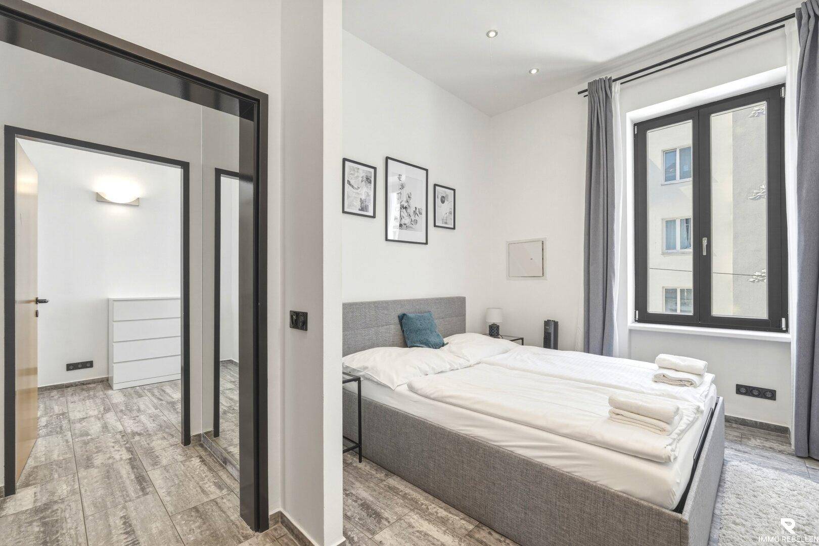 Modernes Schlafzimmer mit grauem Bett und Zugang zu einem weiteren Raum, hell und einladend.