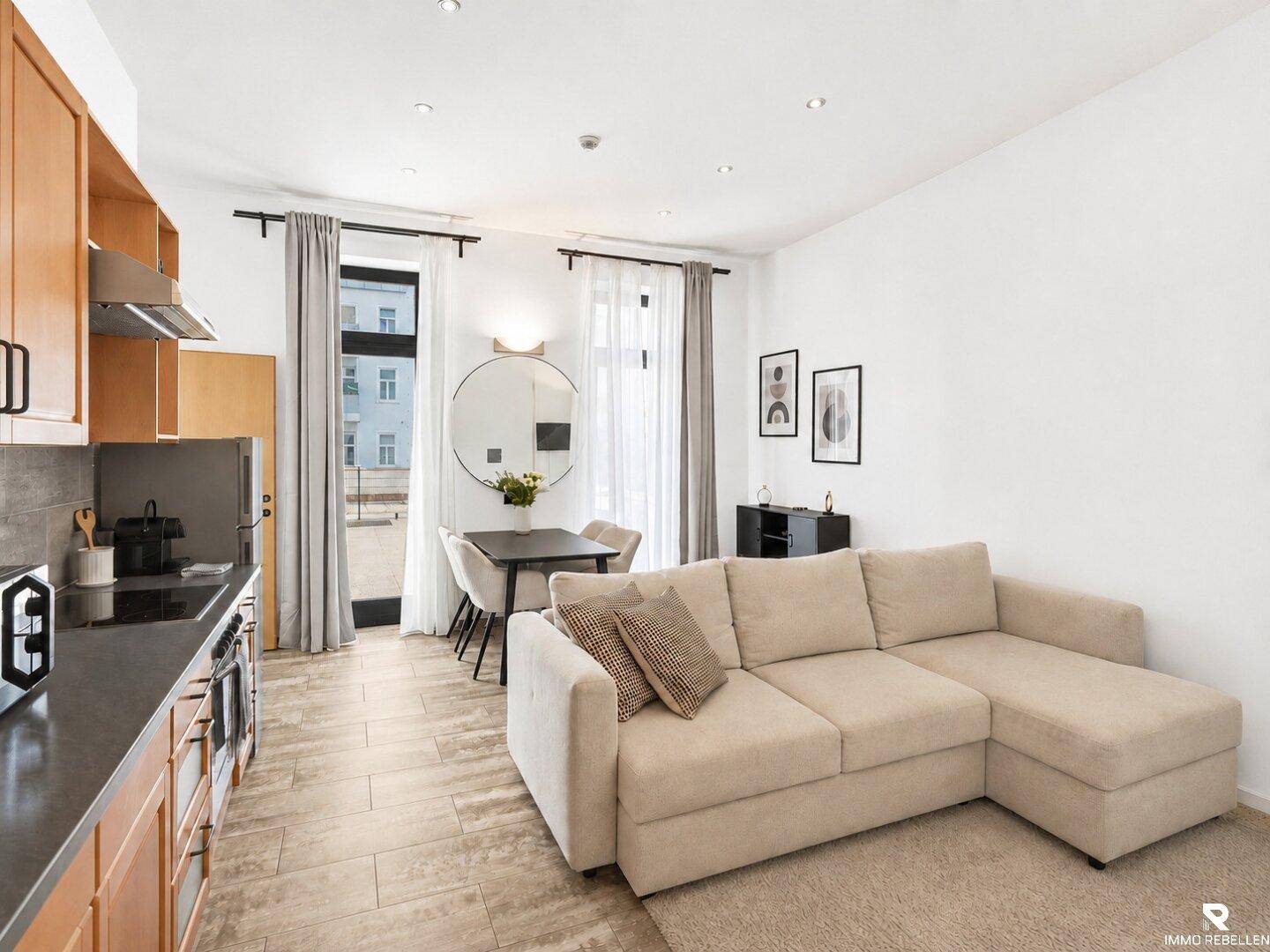 Heller offener Wohn-Essbereich mit integrierter Küche, beigem Sofa und Zugang zu einem Balkon.