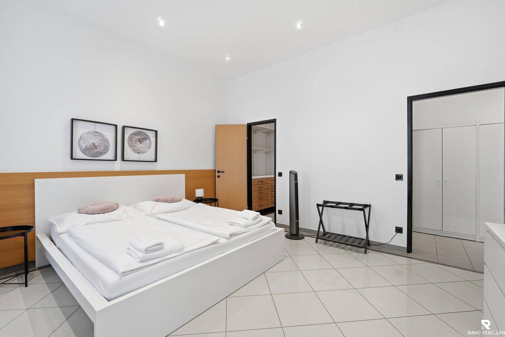 Geräumiges Schlafzimmer mit Doppelbett, weißer Kommode und Zugang zu einem weiteren Raum.