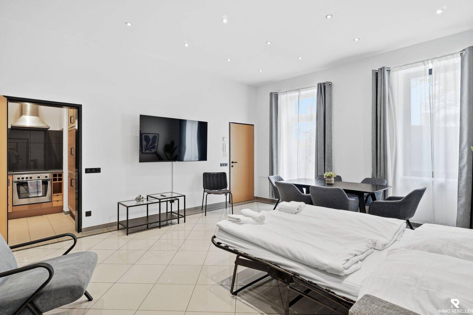 Heller Wohn- und Essbereich mit Schlafsofa, großem Fenster und Blick in eine separate Küche.