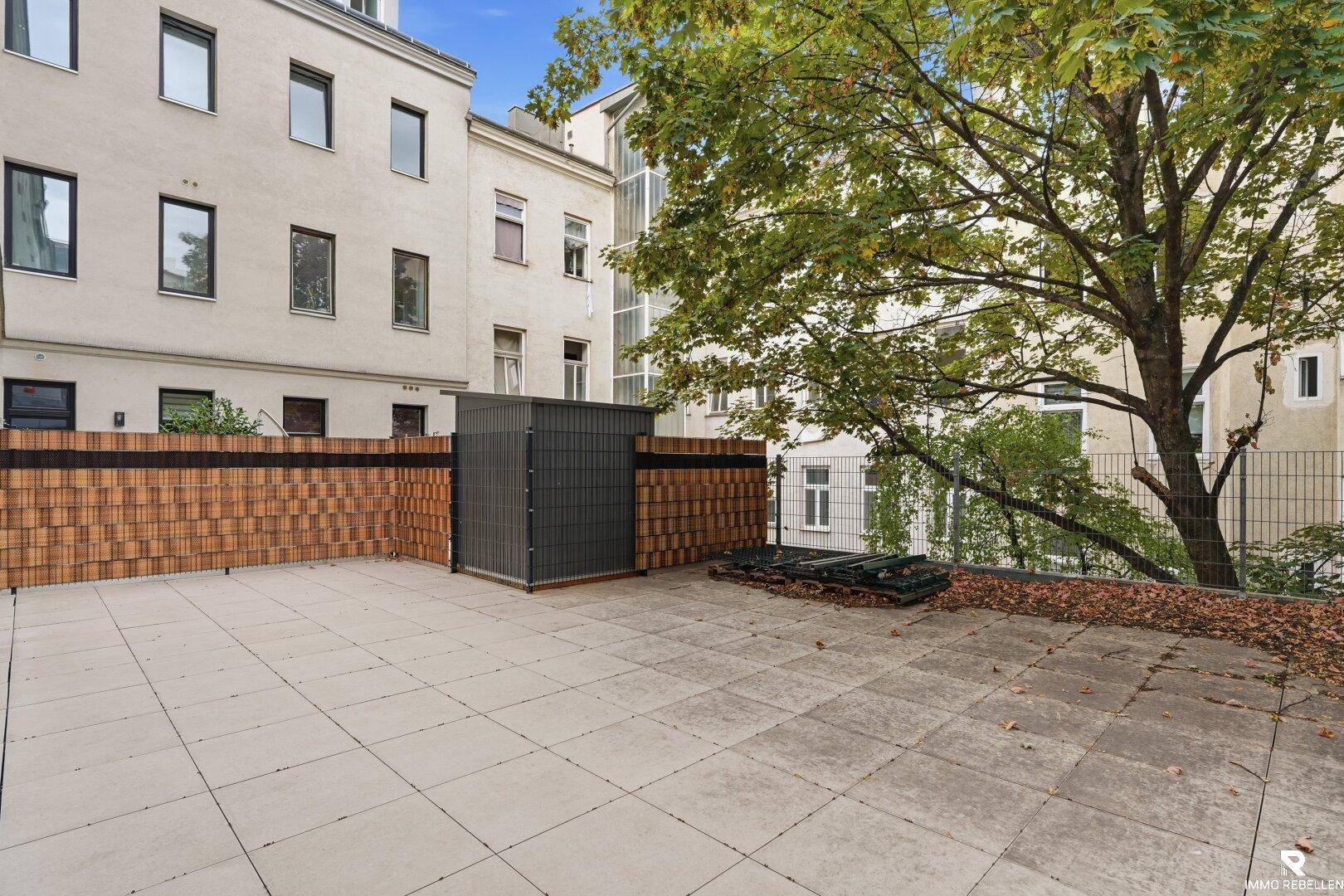 Geräumige, geflieste Terrasse mit einem kleinen Schuppen und Blick auf umliegende Gebäude und Bäume.
