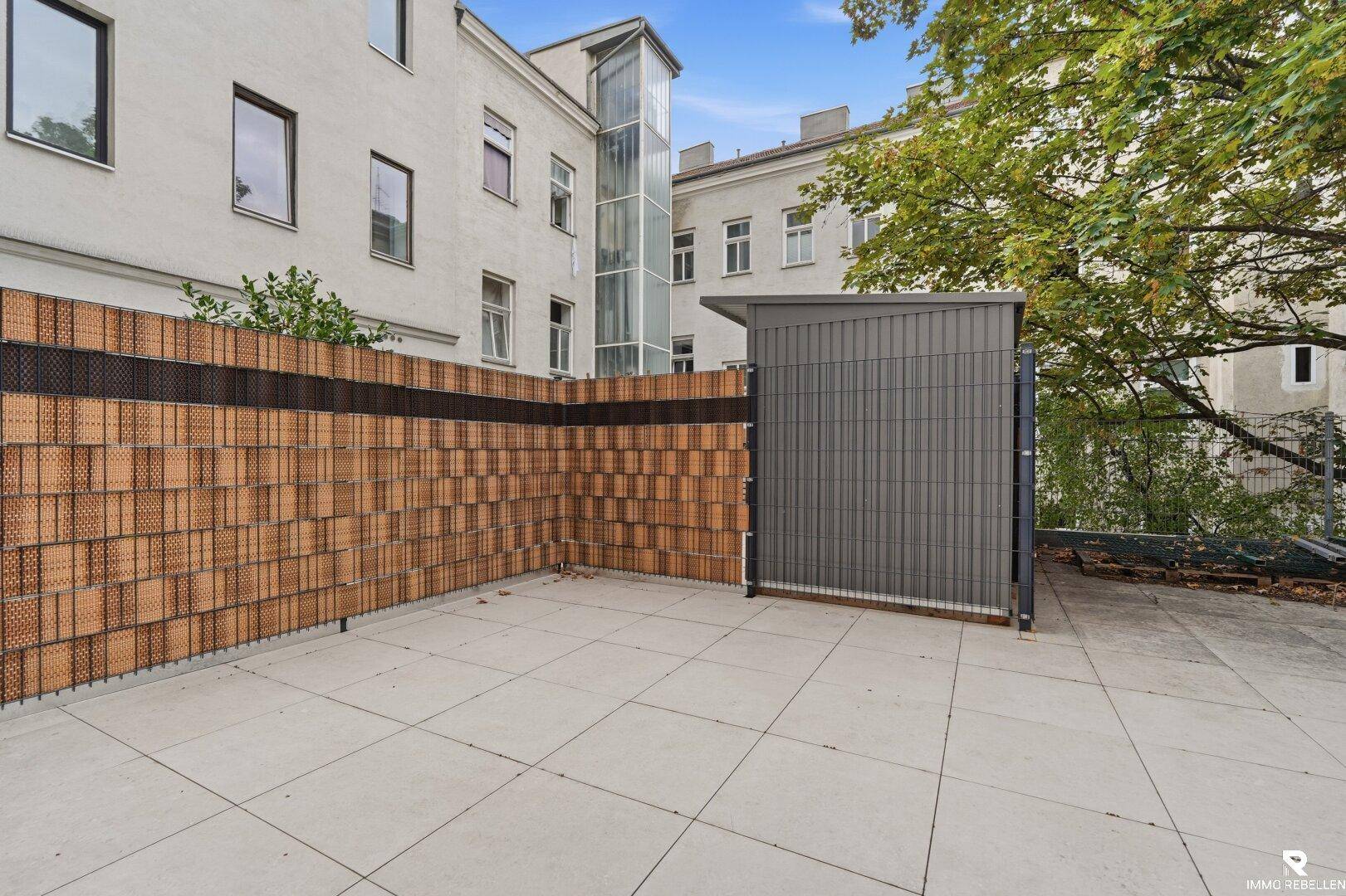 Helle Terrasse mit Fliesenboden und einem modernen Schuppen, im Hintergrund ist ein Aufzug sichtbar.