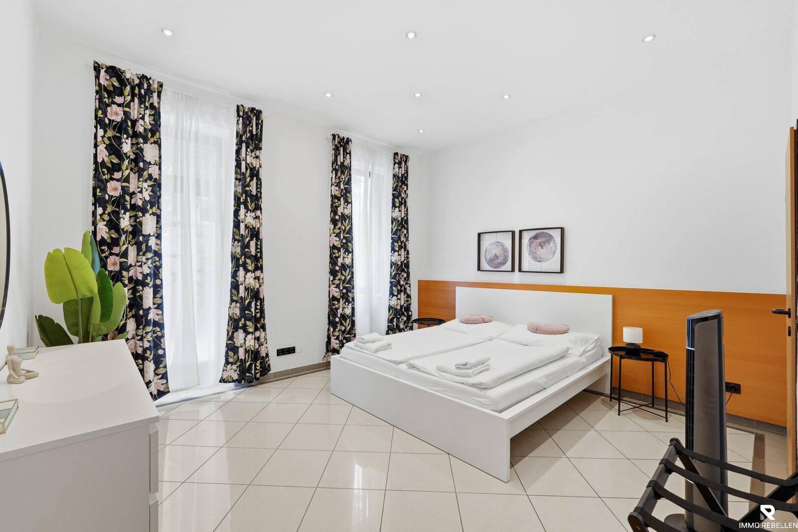 Helles Schlafzimmer mit Doppelbett, floralen Vorhängen und einem modernen Sideboard.
