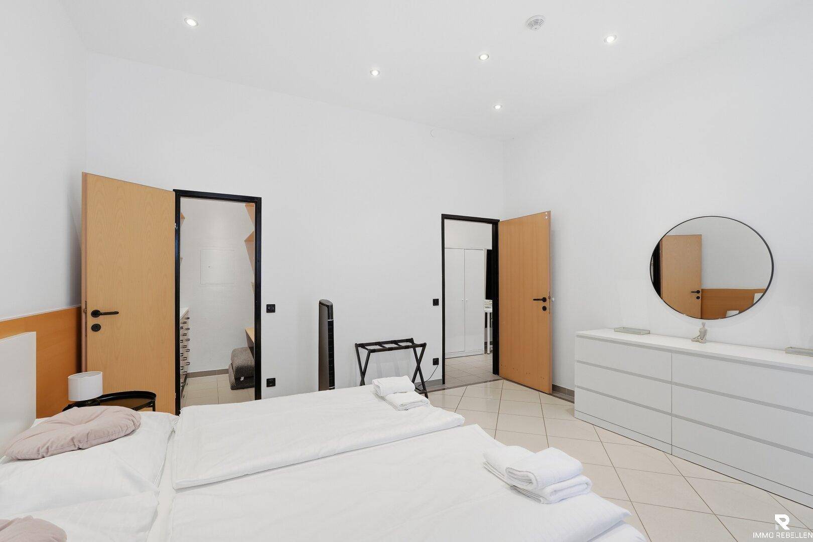 Helles Schlafzimmer mit Doppelbett, weißer Kommode und Zugang zu weiteren Räumen.