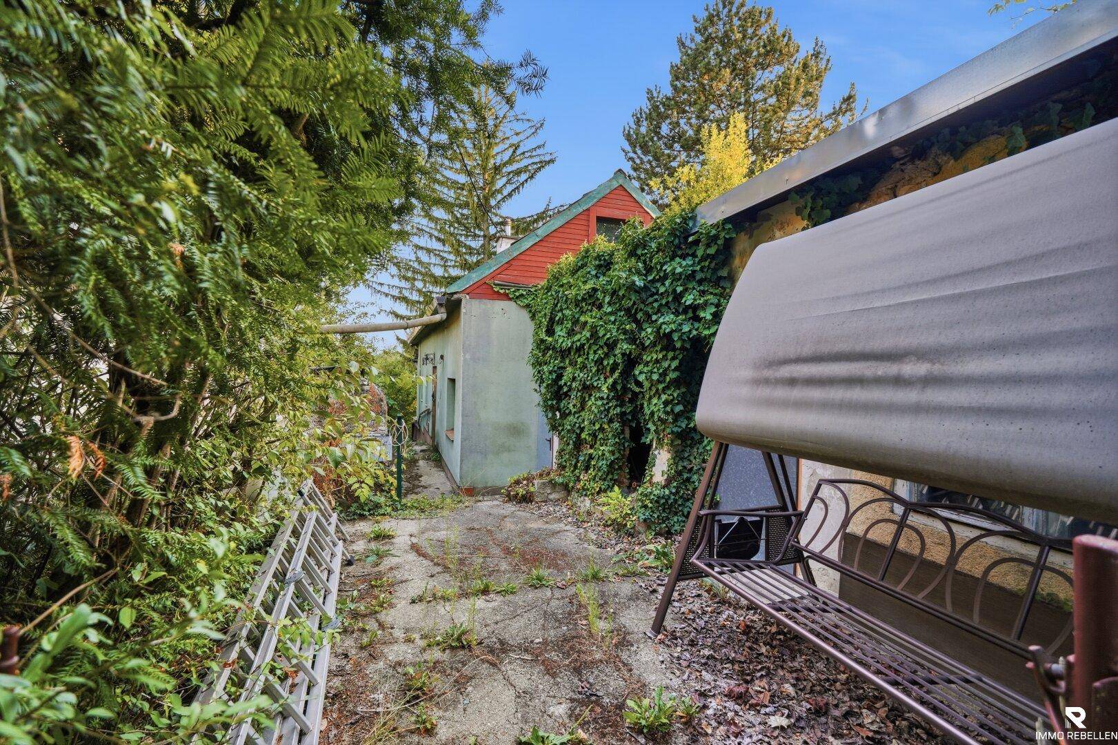 Schmaler Weg neben dem Haus mit einer Hollywoodschaukel und dichtem Bewuchs.